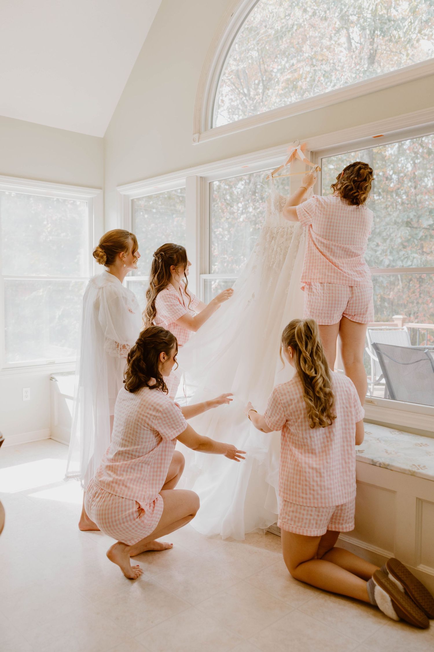 Bridesmaids in pink robes helping arrange a white wedding gown by a bright window.
