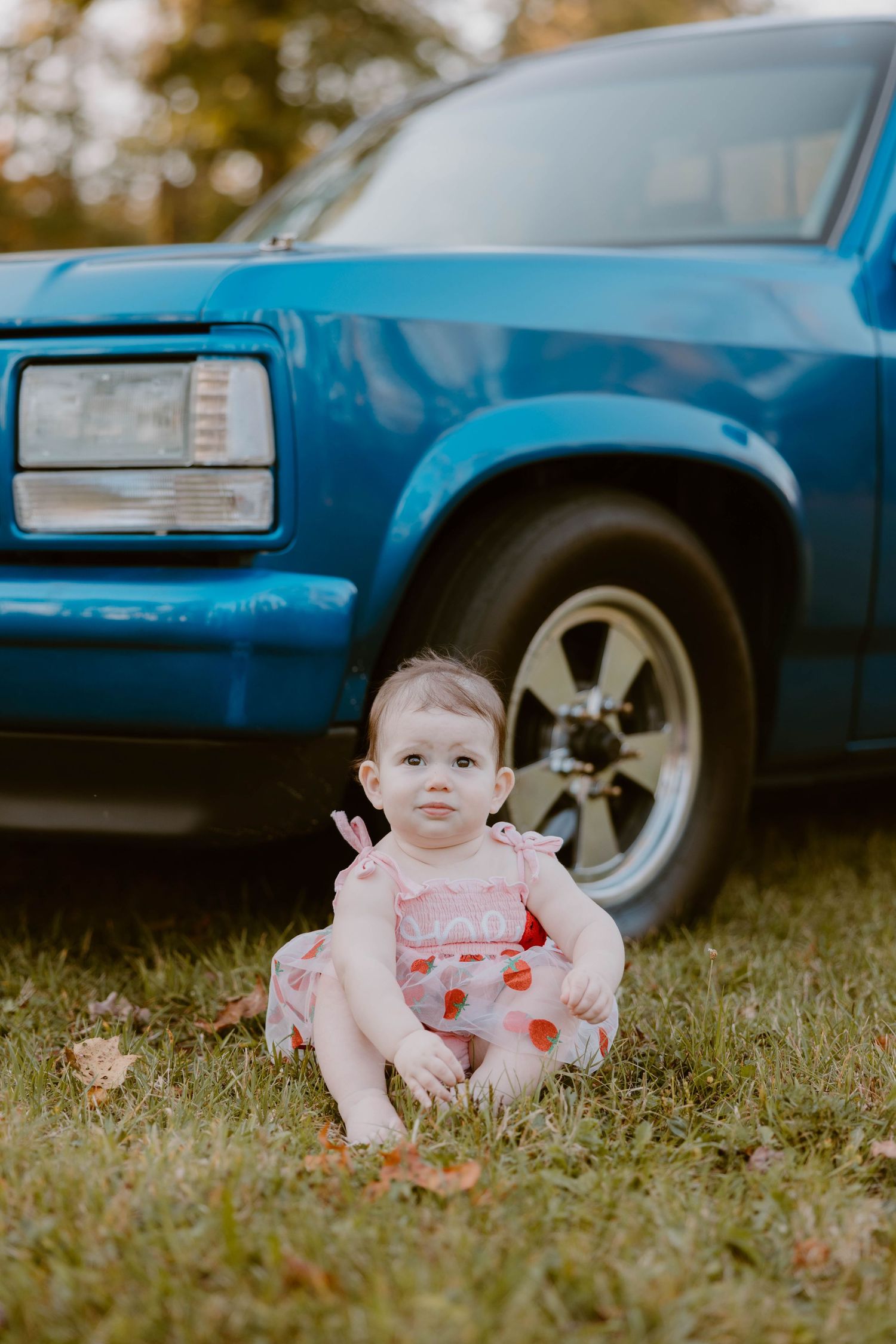 Baby girl in pink dress sits in grass beside a classic blue vintage truck outdoors.