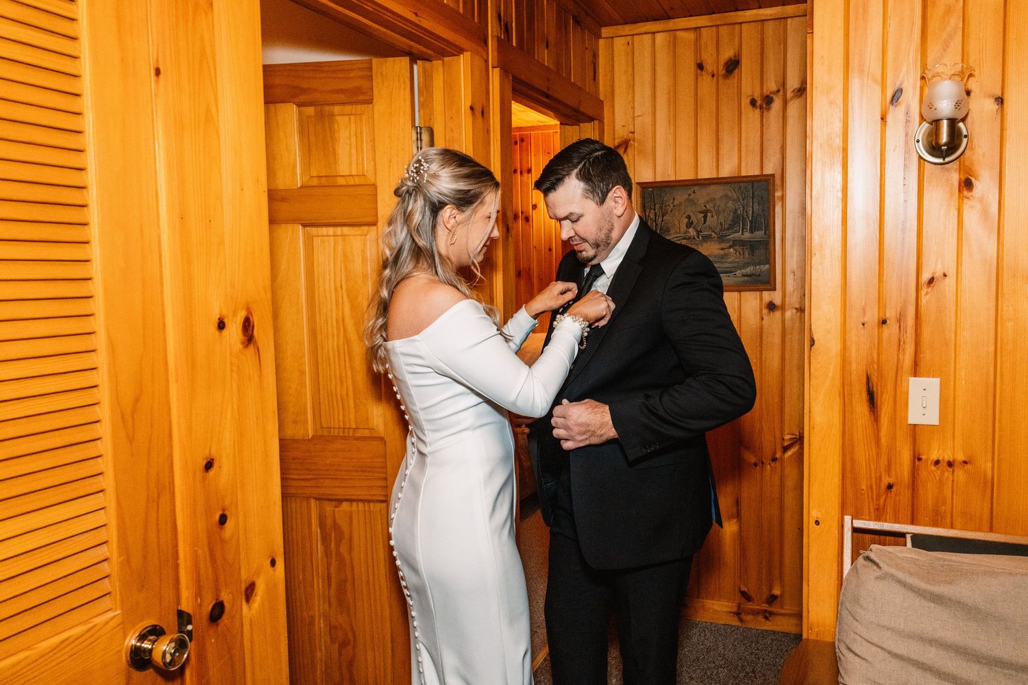 Bride adjusts groom's boutonniere in a warm wood-paneled room before their wedding ceremony begins.
