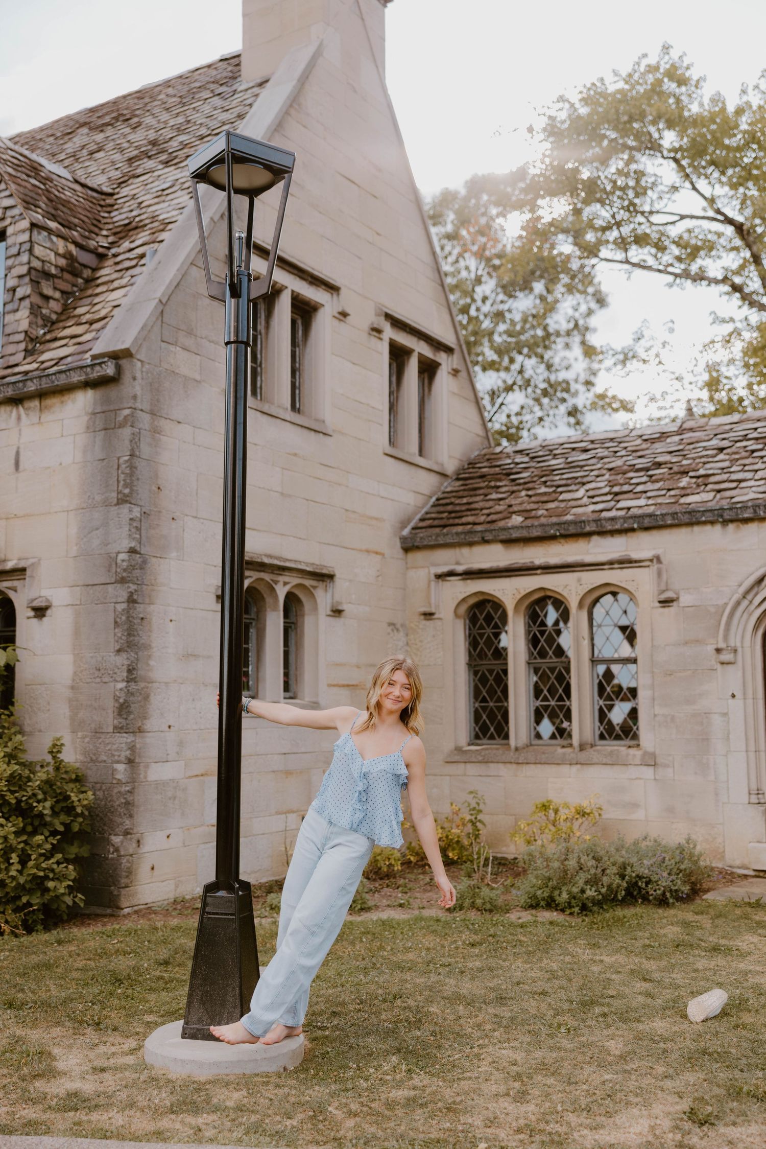 Woman in light blue jumpsuit posing playfully against a black lamppost outside a stone manor.