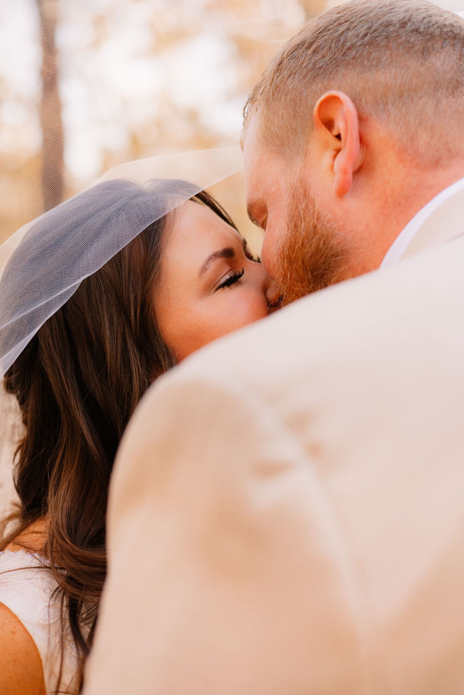 Close-up of bride in veil and groom sharing a romantic kiss outdoors with soft golden bokeh background.
