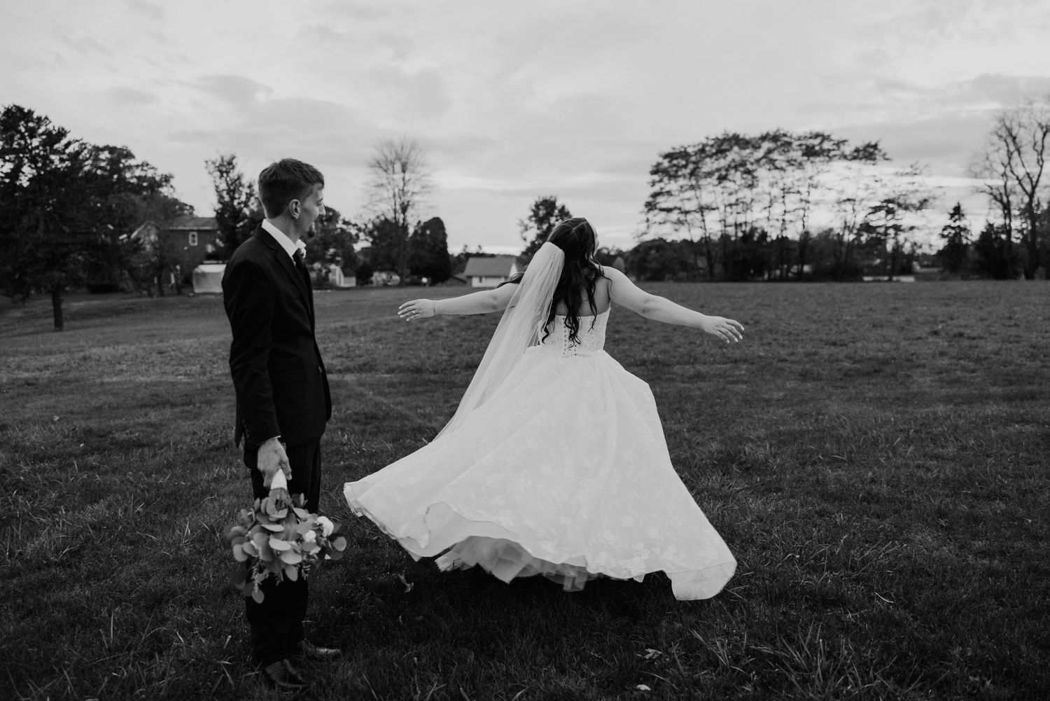 Black and white photo of bride twirling wedding dress in open field at dusk.