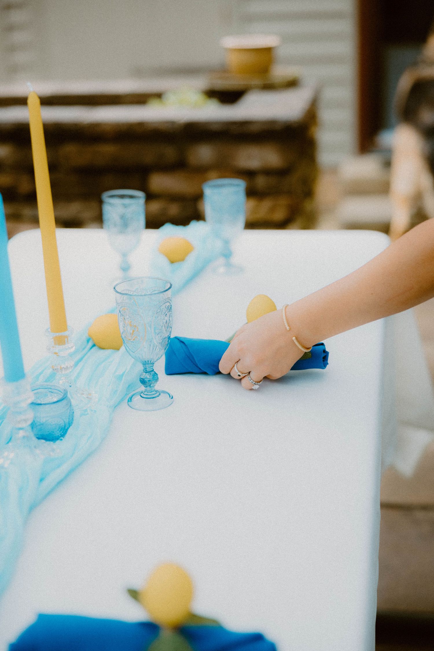 Hand arranging a blue napkin on a white table decorated with lemons, blue glasses, and candles.