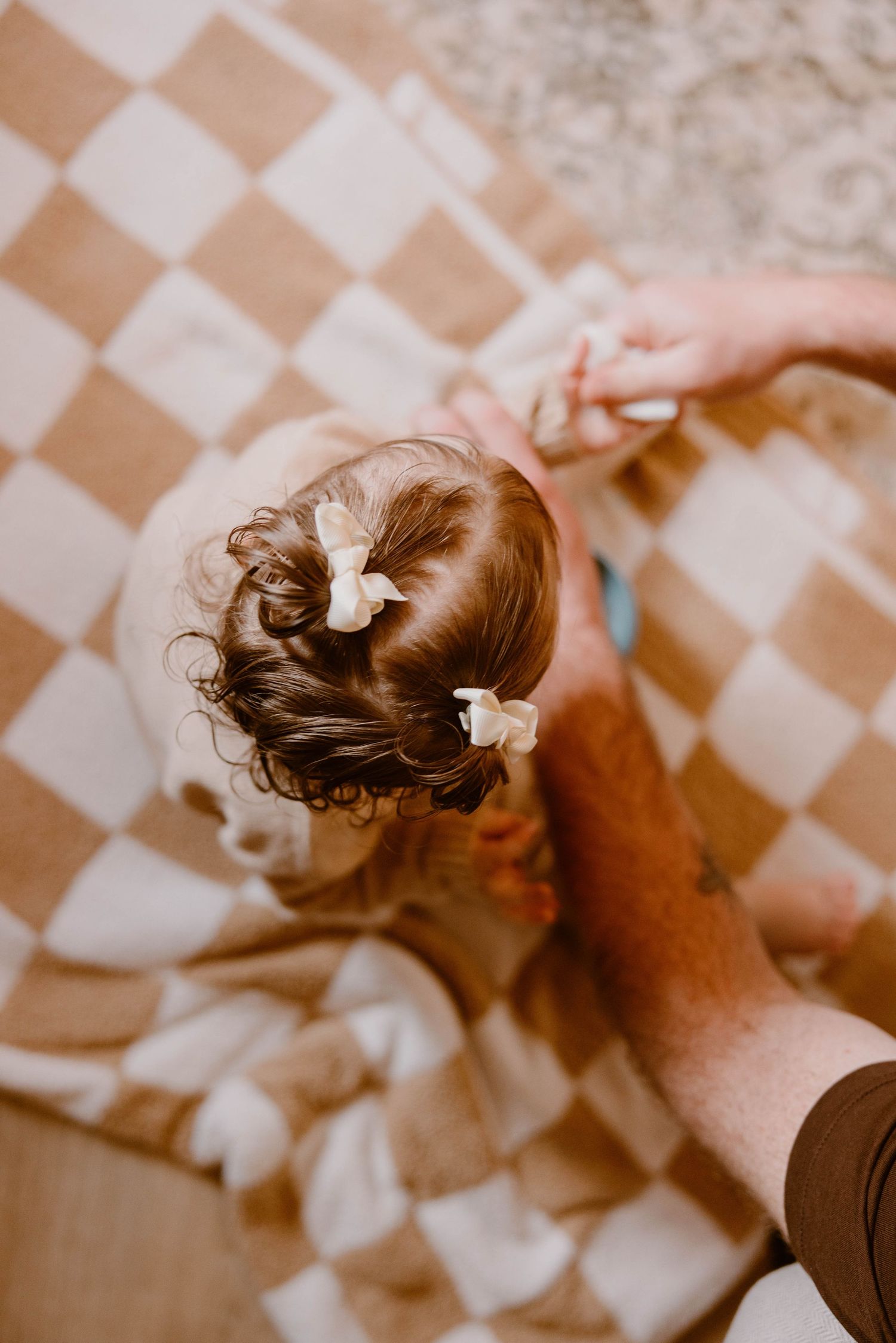 Overhead view of a toddler girl with hair bows sitting on a checkered blanket holding an adults hand.