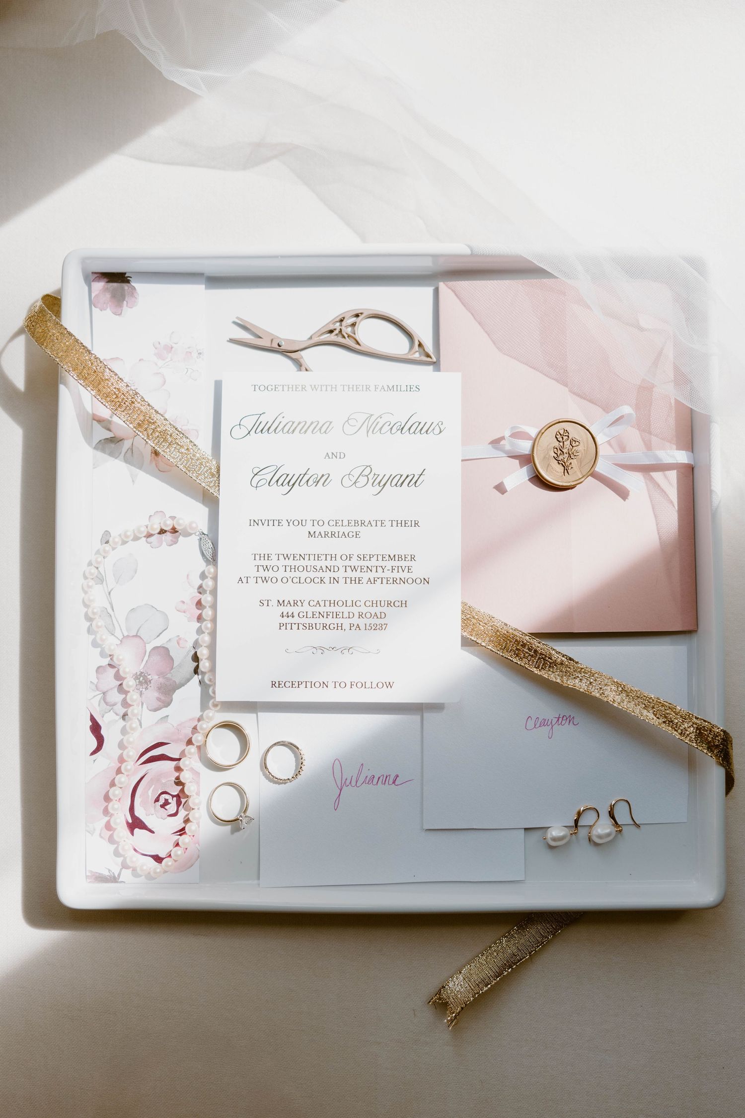 Wedding invitation suite with rings, pearls, and gold ribbon on a white tray.