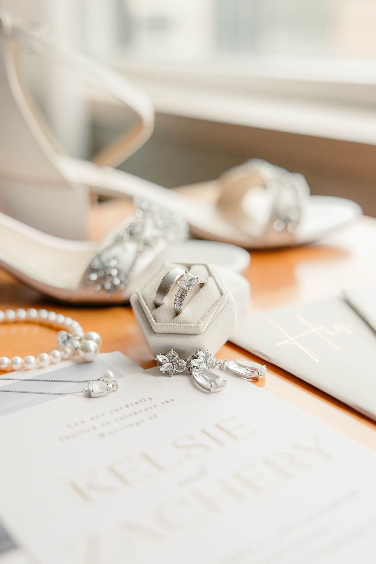 Elegant wedding flat lay featuring rings, pearl bracelet, crystal earrings, heels, and invitation.