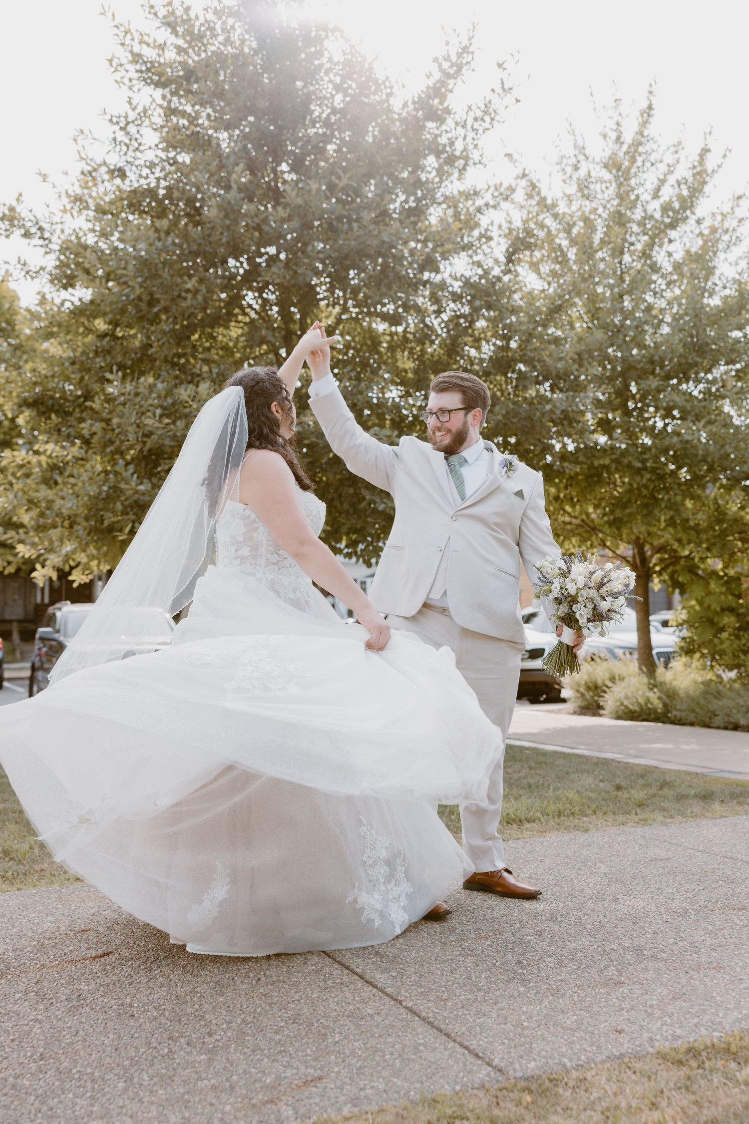 Bride and groom dancing outdoors, groom spinning bride in flowing white wedding dress and veil on sunny day.