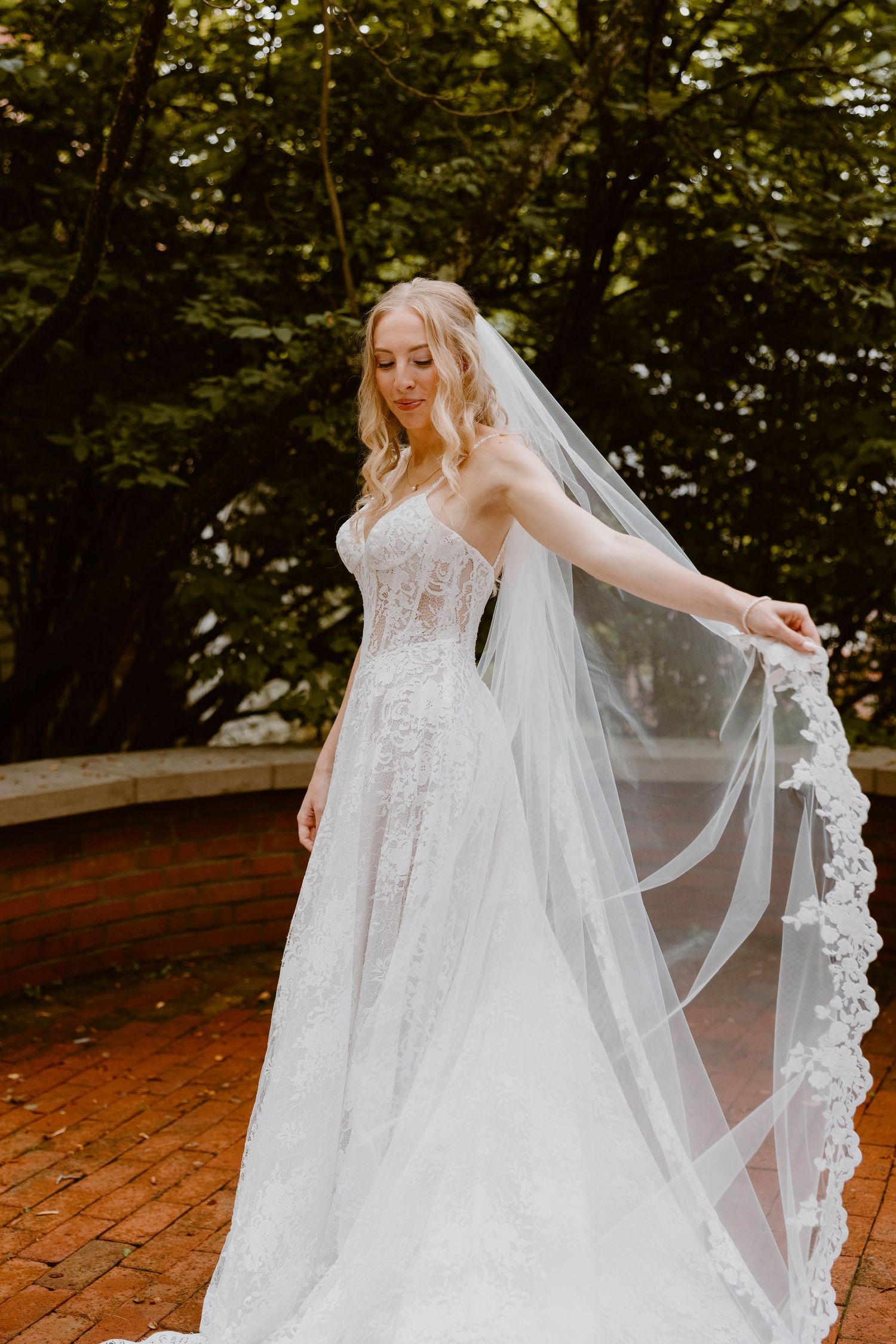 Elegant bride in lace A-line gown holding flowing floral-edged cathedral veil in lush garden setting.