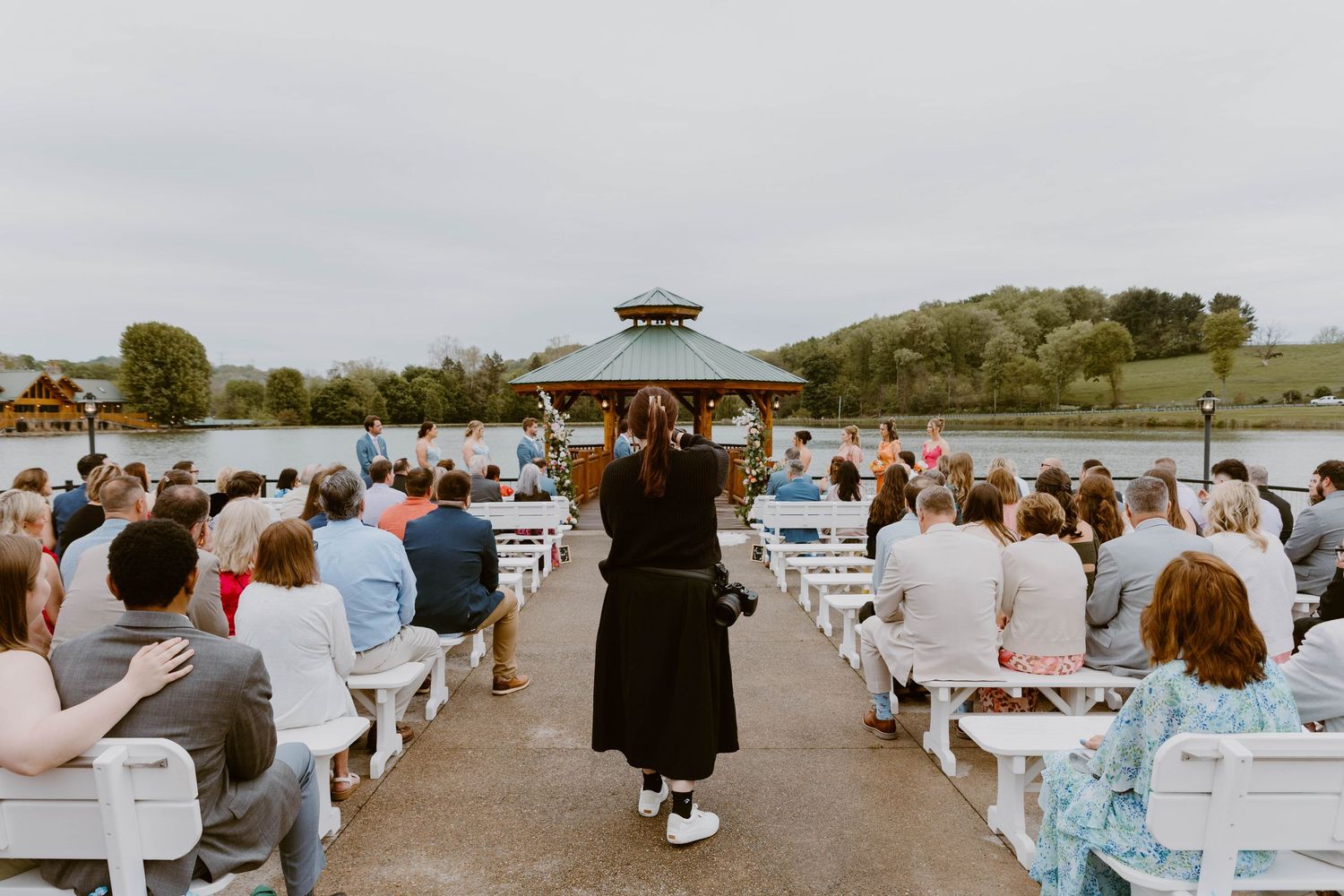 Guests seated on white benches at an outdoor lakeside wedding ceremony with a gazebo backdrop.