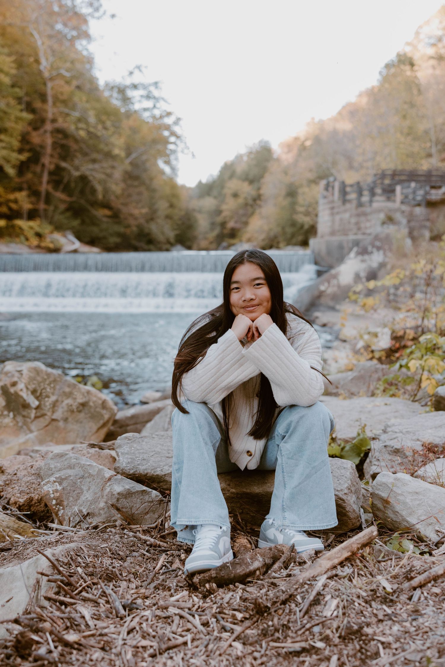 Young woman in white sweater and jeans sits on rocks by a waterfall in autumn forest.