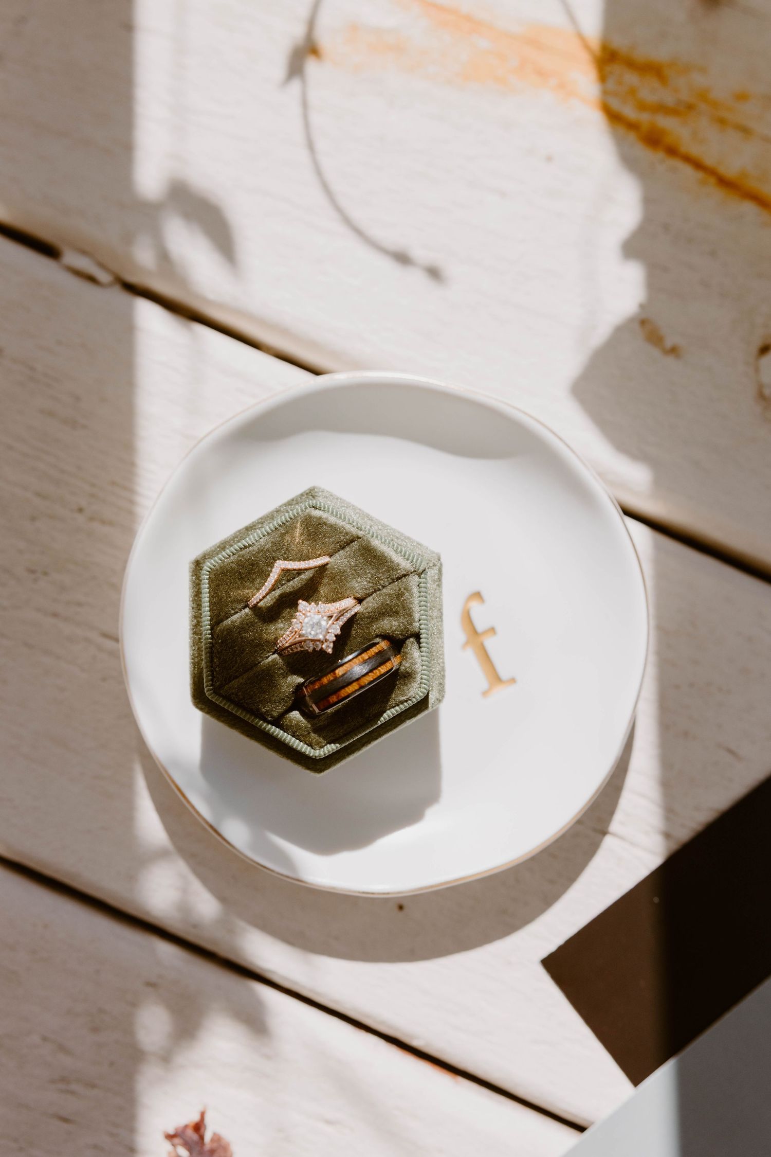 Diamond engagement ring and gold wedding bands nestled in a sage green velvet hexagon ring box on a white plate.