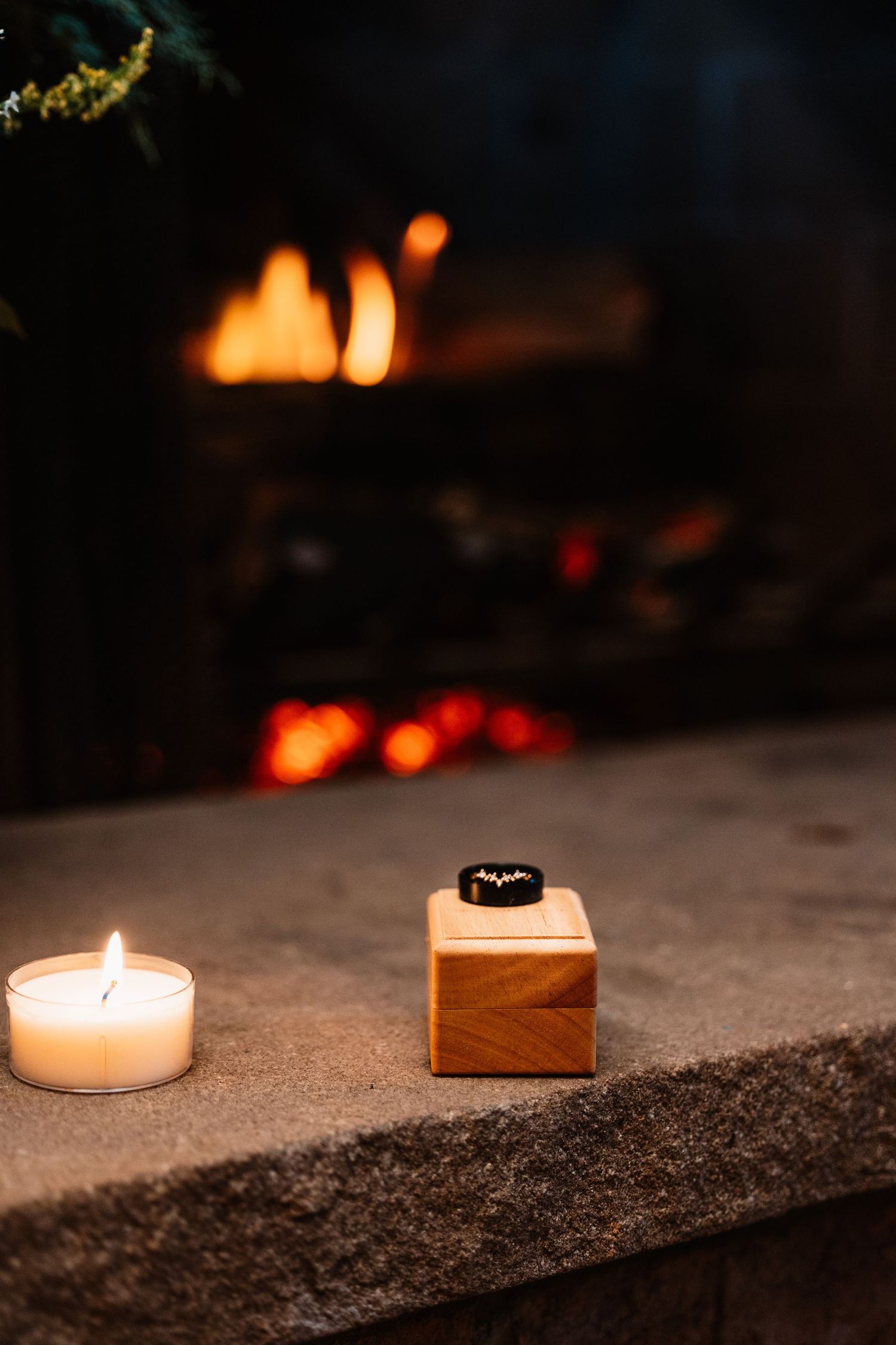 Wooden ring box with wedding band beside glowing candle near romantic fireplace ambiance.