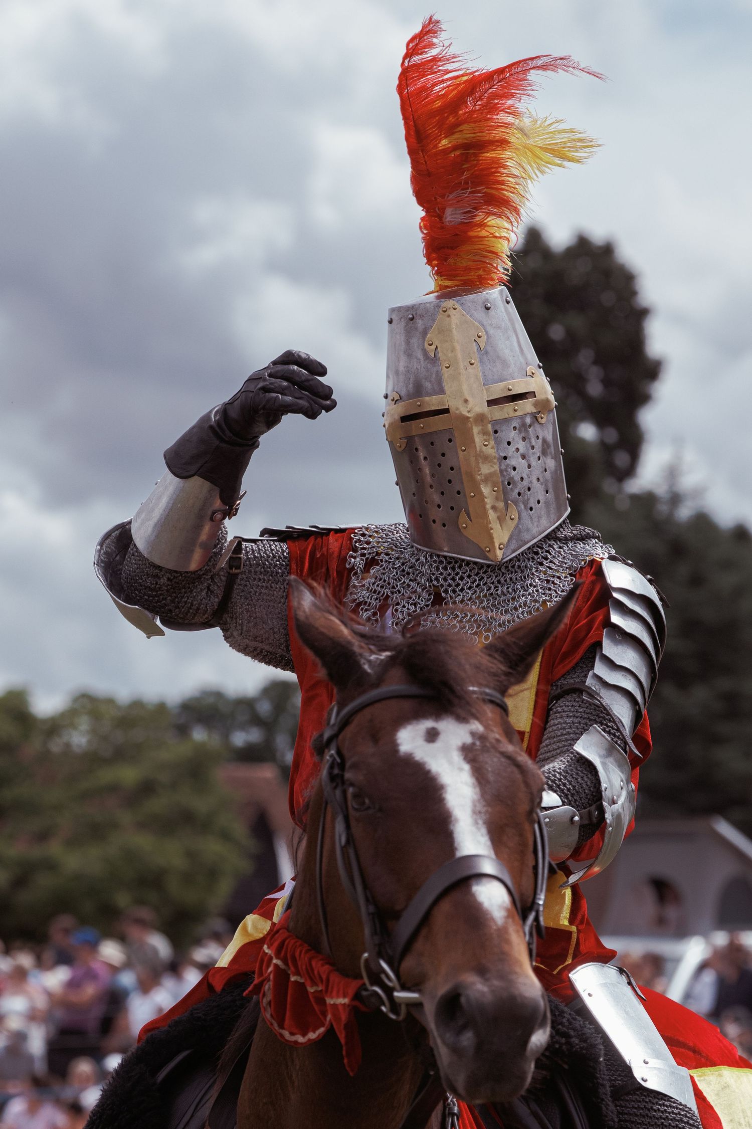 Knight in full armor with red feathered helmet rides brown horse at medieval jousting reenactment event.