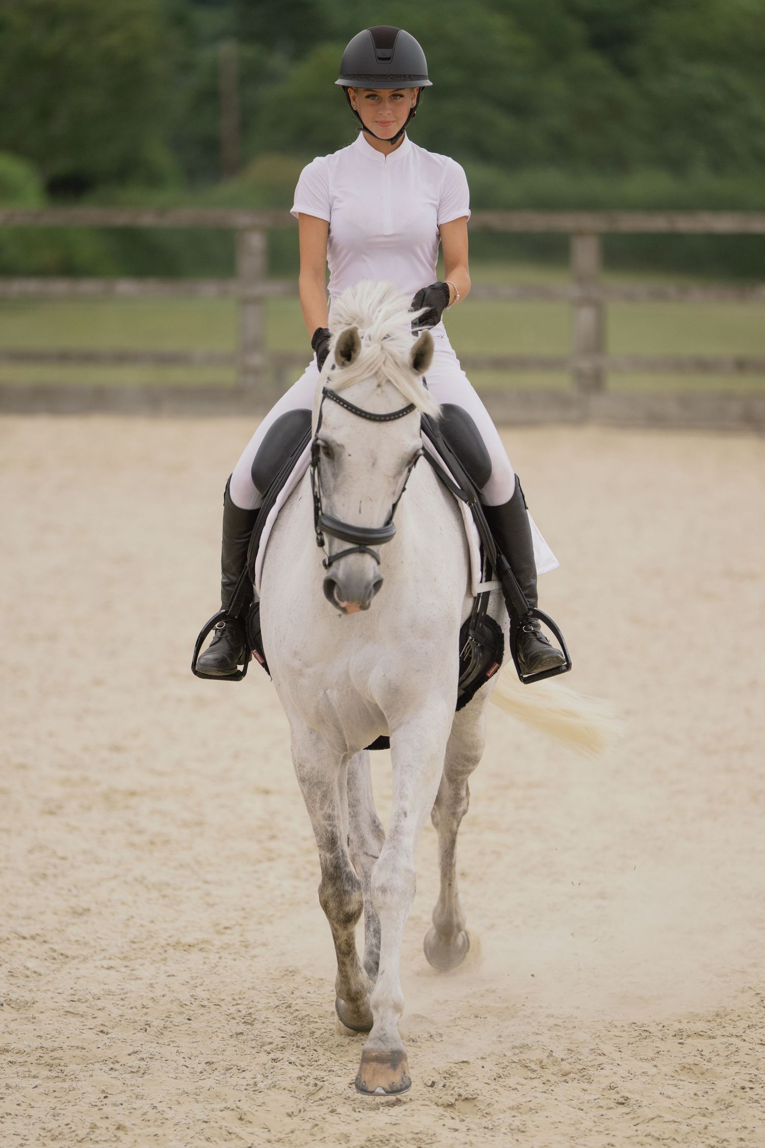 Young female equestrian in white top and helmet riding a grey horse in a sandy arena.