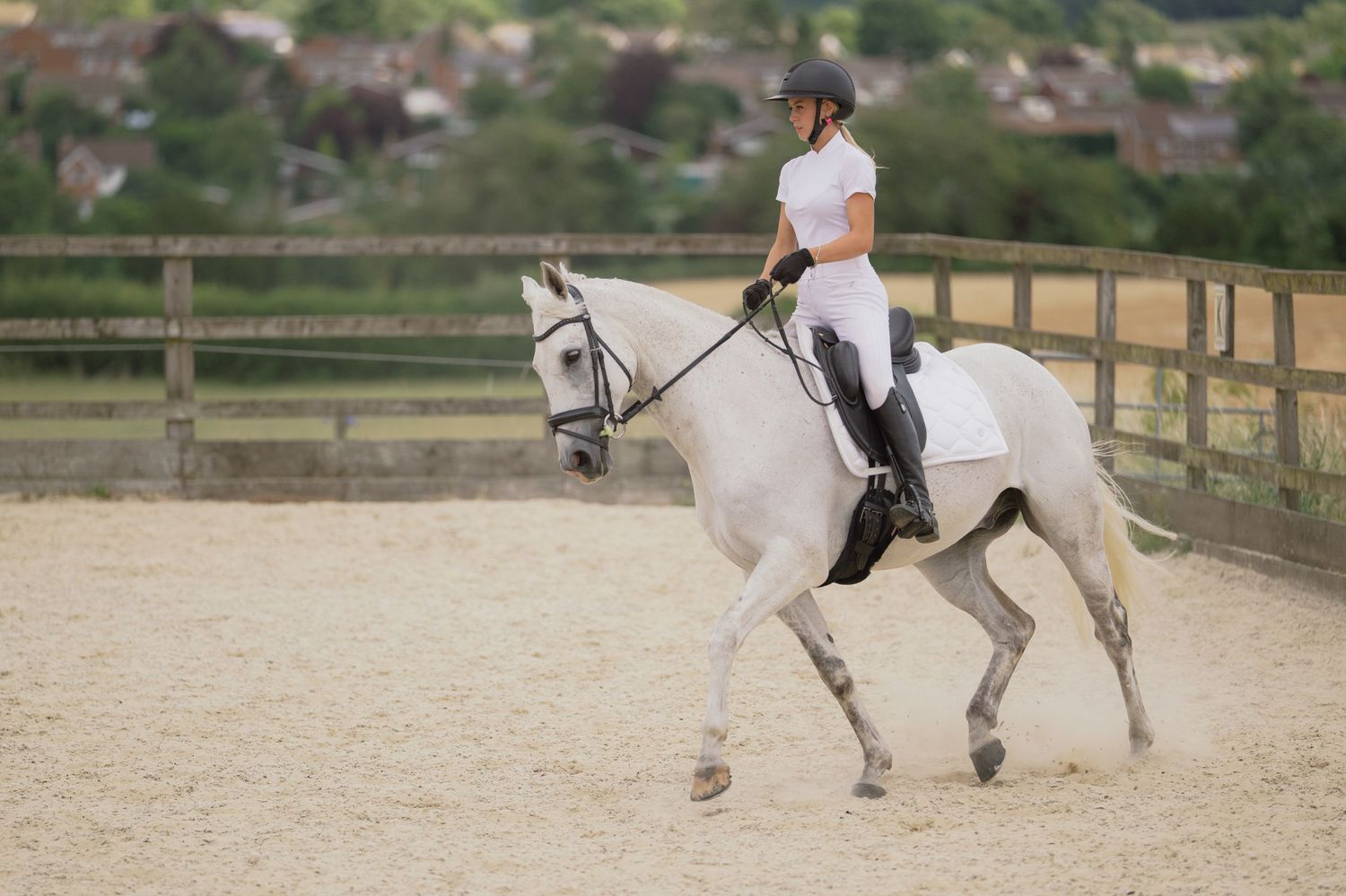 A woman in white riding attire practices dressage on a grey horse in a sandy outdoor arena.