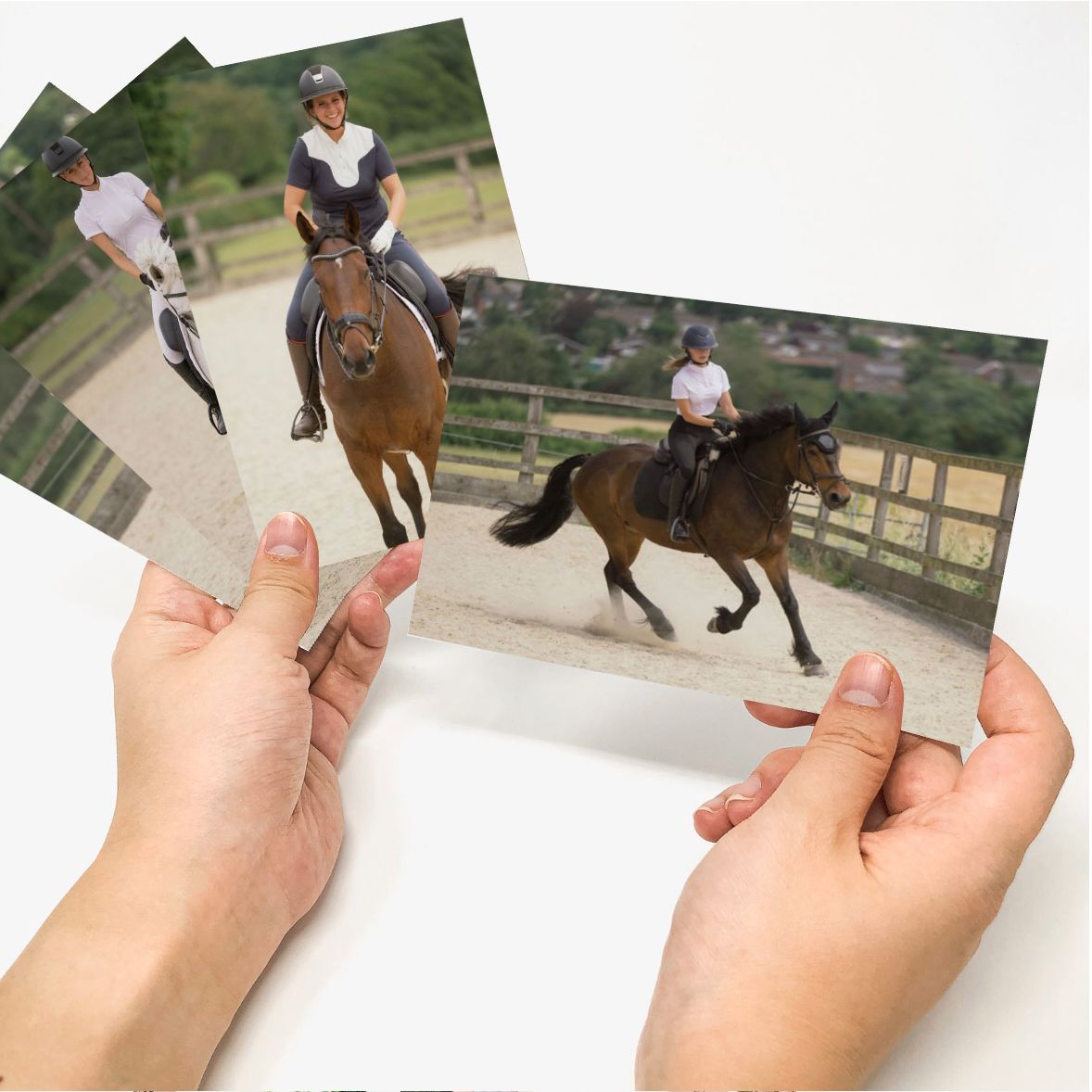 Hands holding printed photos of a person riding a brown horse in an equestrian arena during a dressage session.