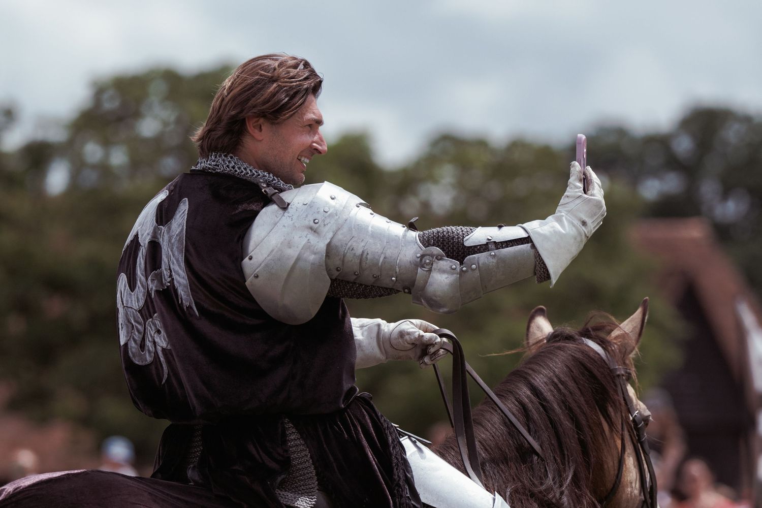 A medieval knight in armor rides a horse, raising his hand in a triumphant gesture at an outdoor event.