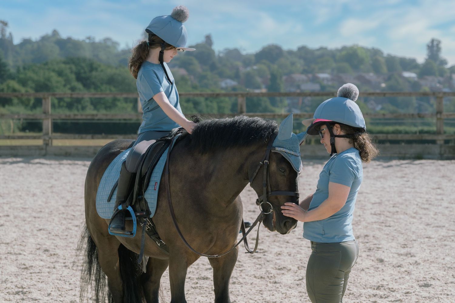 Young girl riding a dark horse during a riding lesson with an instructor in an outdoor arena.