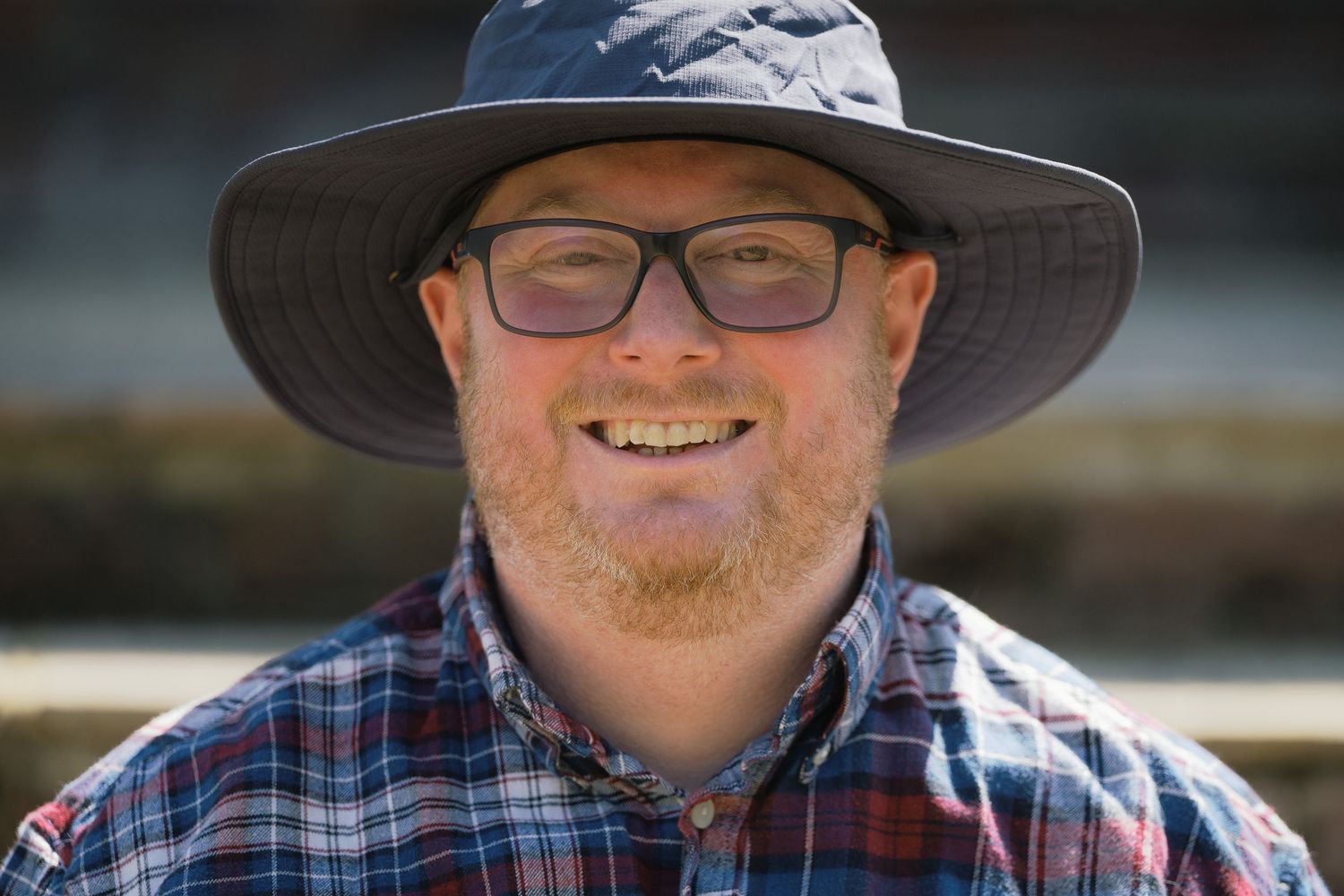 Smiling man with red beard wearing glasses and a wide-brim hat and plaid shirt outdoors.