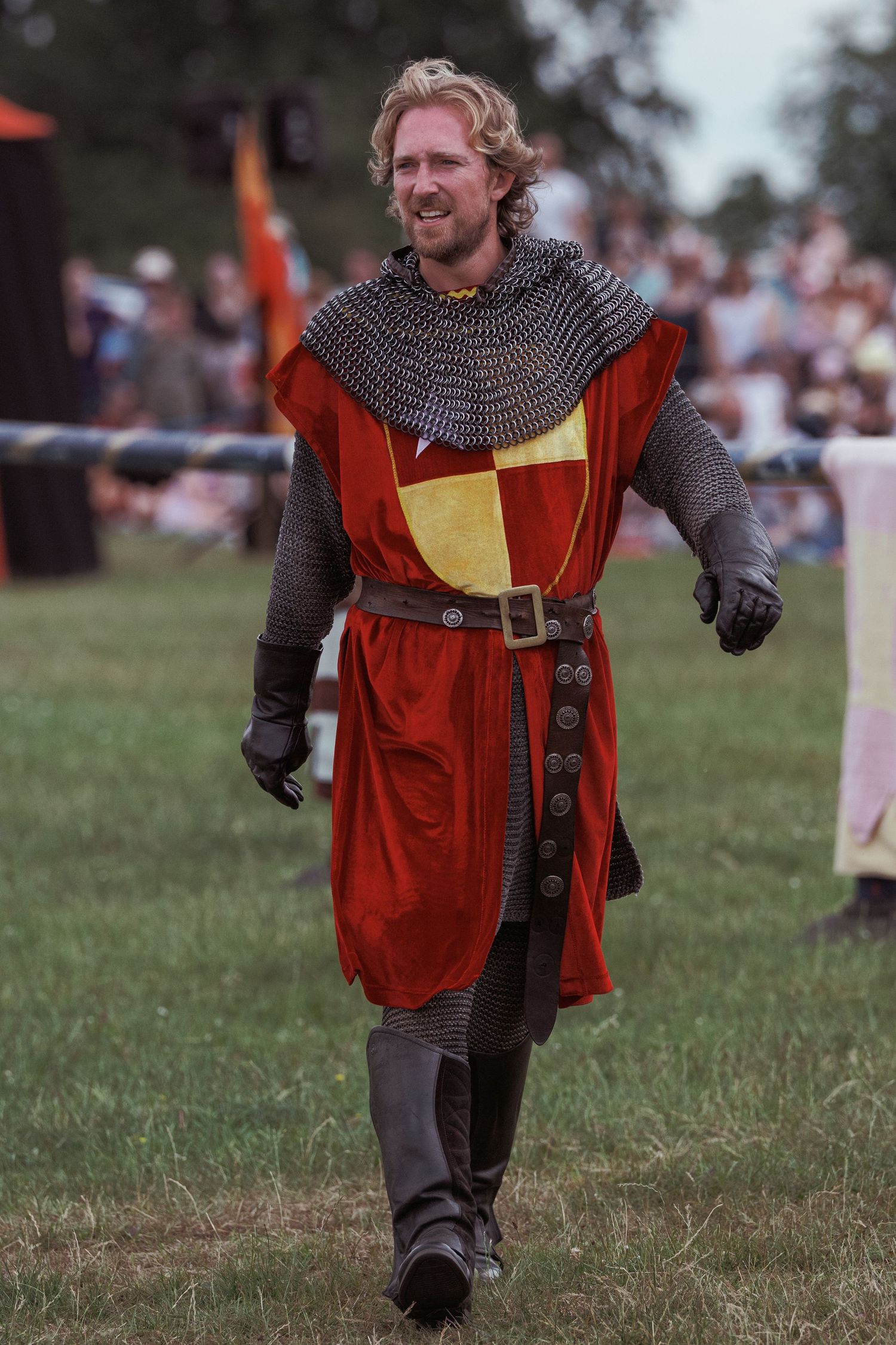A smiling man in medieval knight costume with chainmail and red-gold tunic walks across green grass at a Renaissance fair.