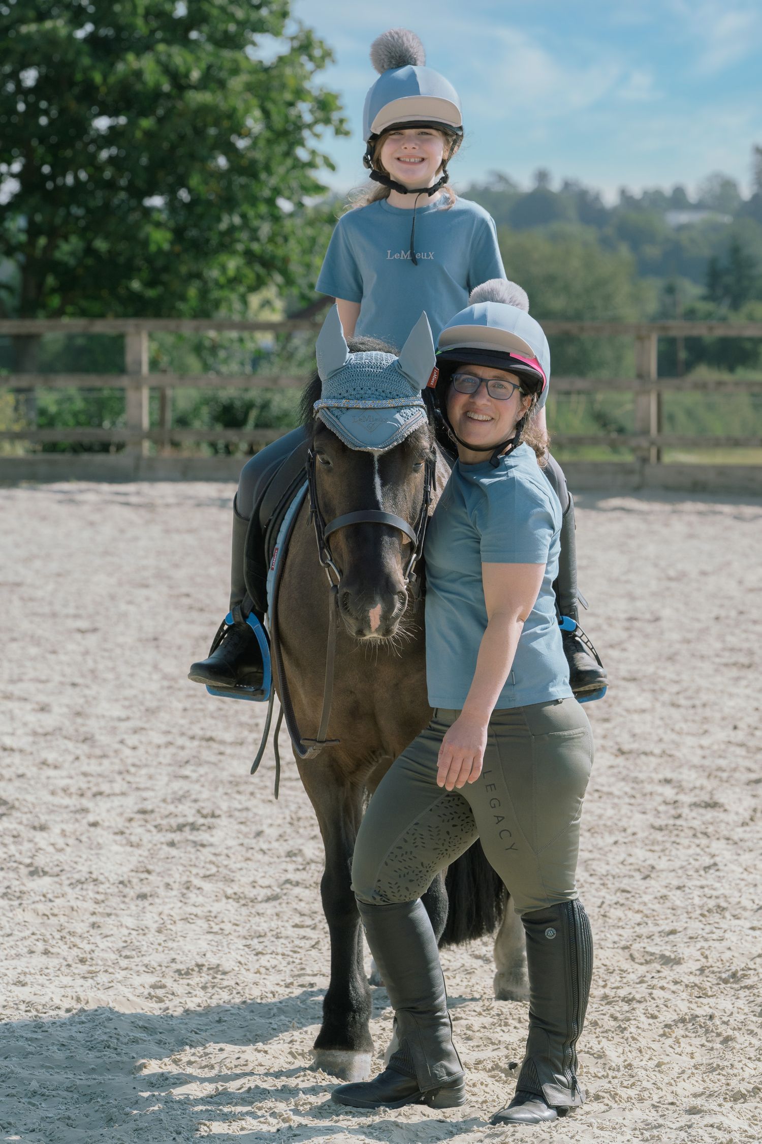 Woman and child in riding helmets pose with a dark pony in a sandy equestrian arena on a sunny day.