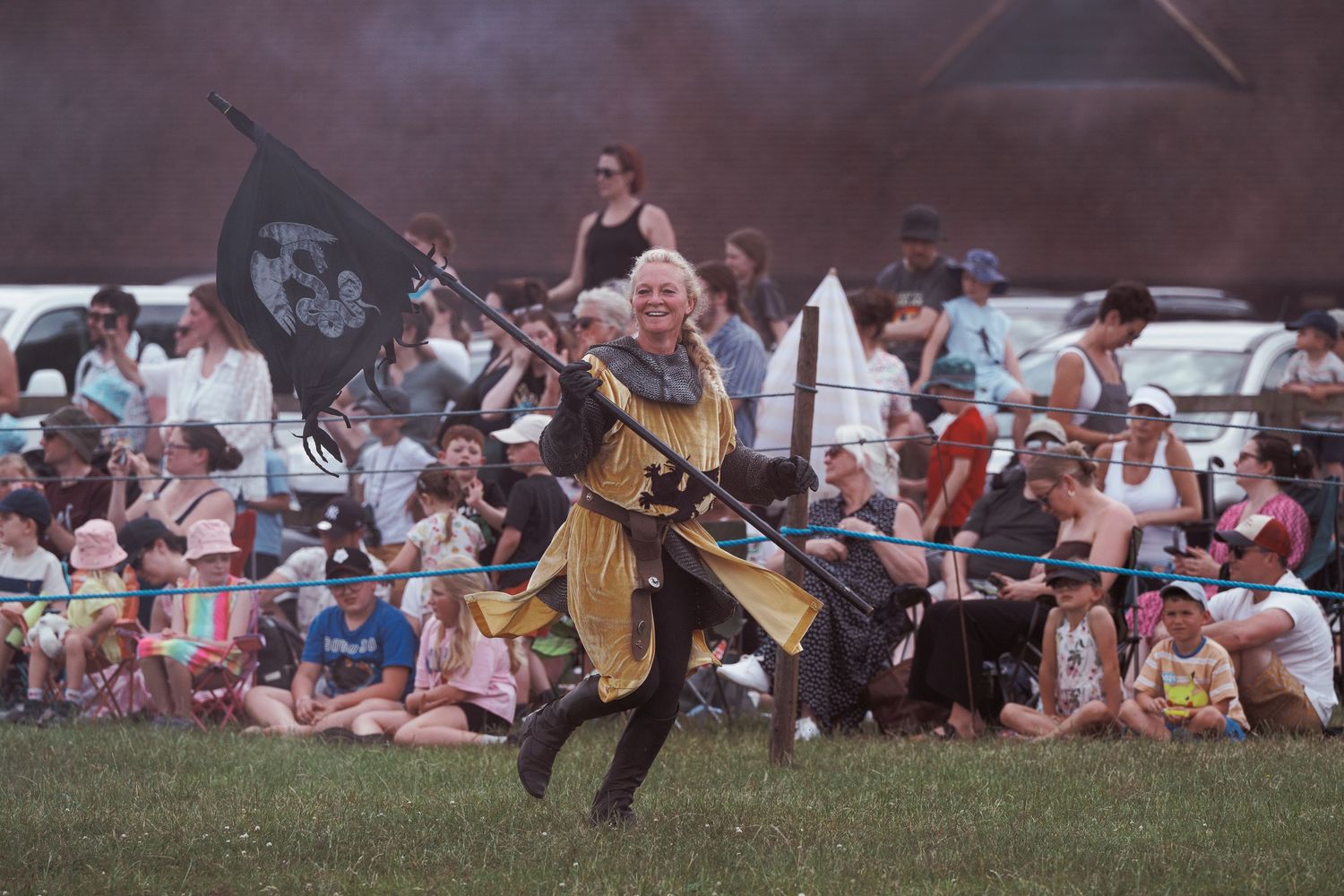 Medieval performer in golden costume carries pirate flag at Renaissance faire while crowd watches on grassy field.