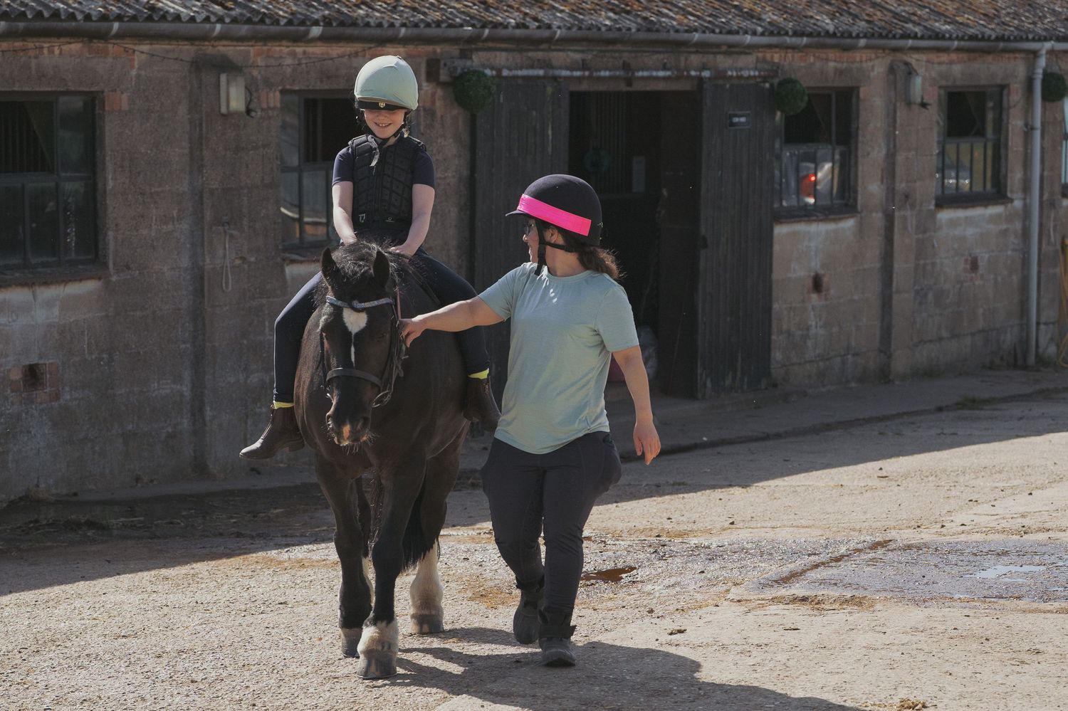 A young rider in a helmet sits atop a dark pony led by an instructor in a pink visor at a stable yard.