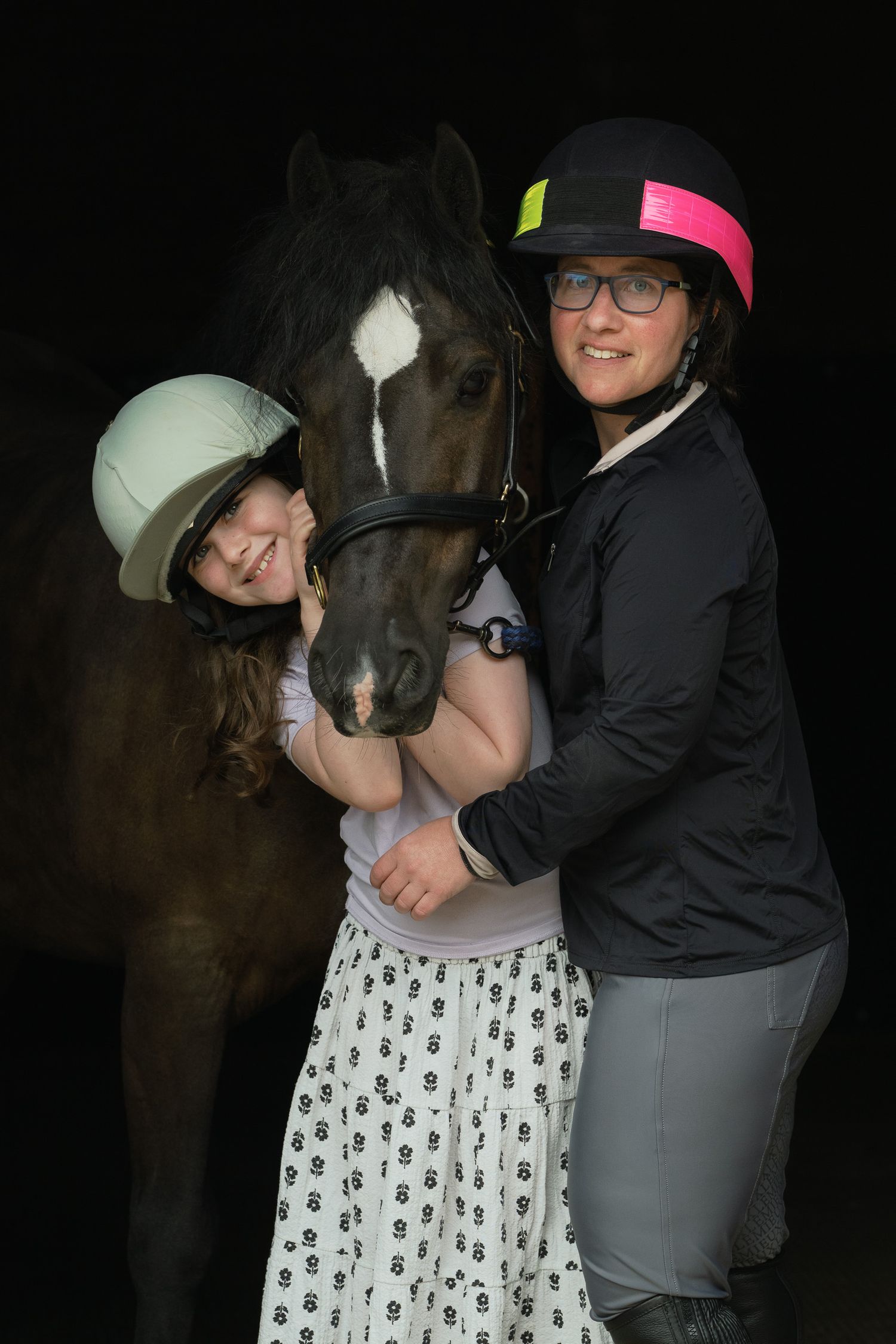 Two people wearing riding helmets affectionately pose with a dark horse against a black background.