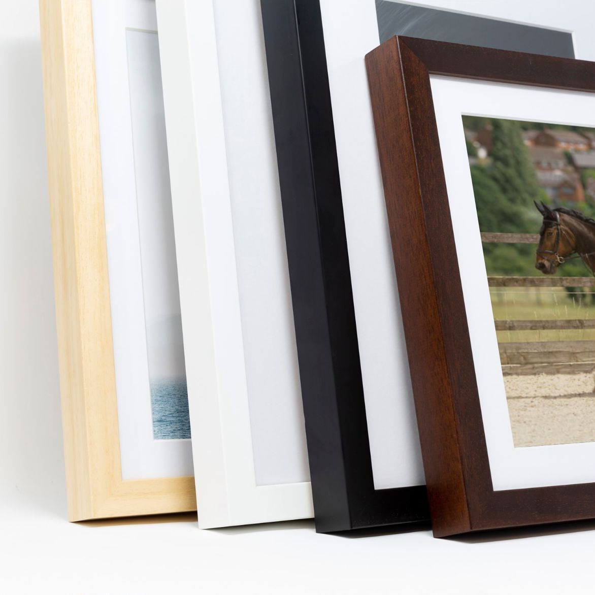Four picture frames in natural wood, white, black, and dark brown finishes leaning against a white wall displaying photos.