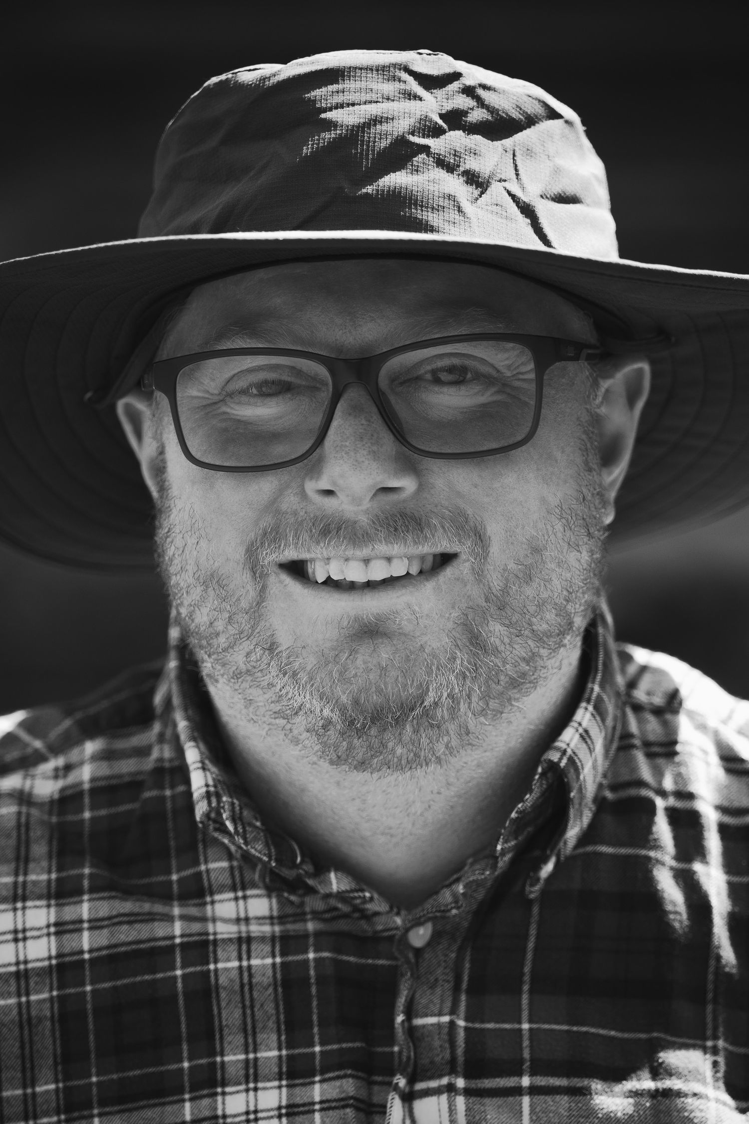 Black and white portrait of a smiling middle-aged man with beard, glasses, and wide-brim hat wearing a plaid shirt.