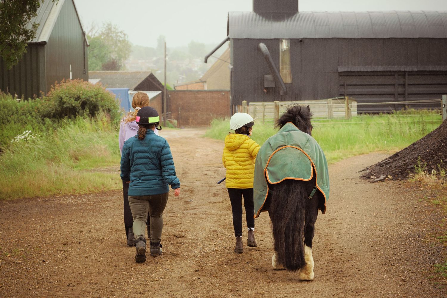 Two children and a pony walking along a farm path toward rustic barns on a rainy day.