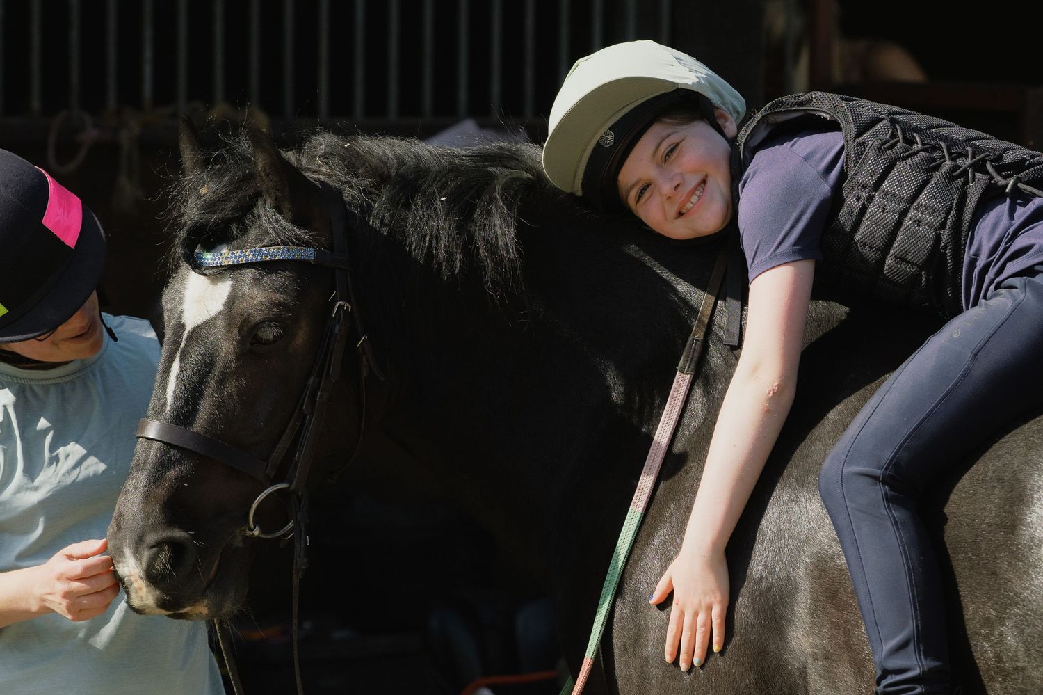 Child in helmet joyfully hugs black horse while riding, assisted by instructor during equestrian therapy session.