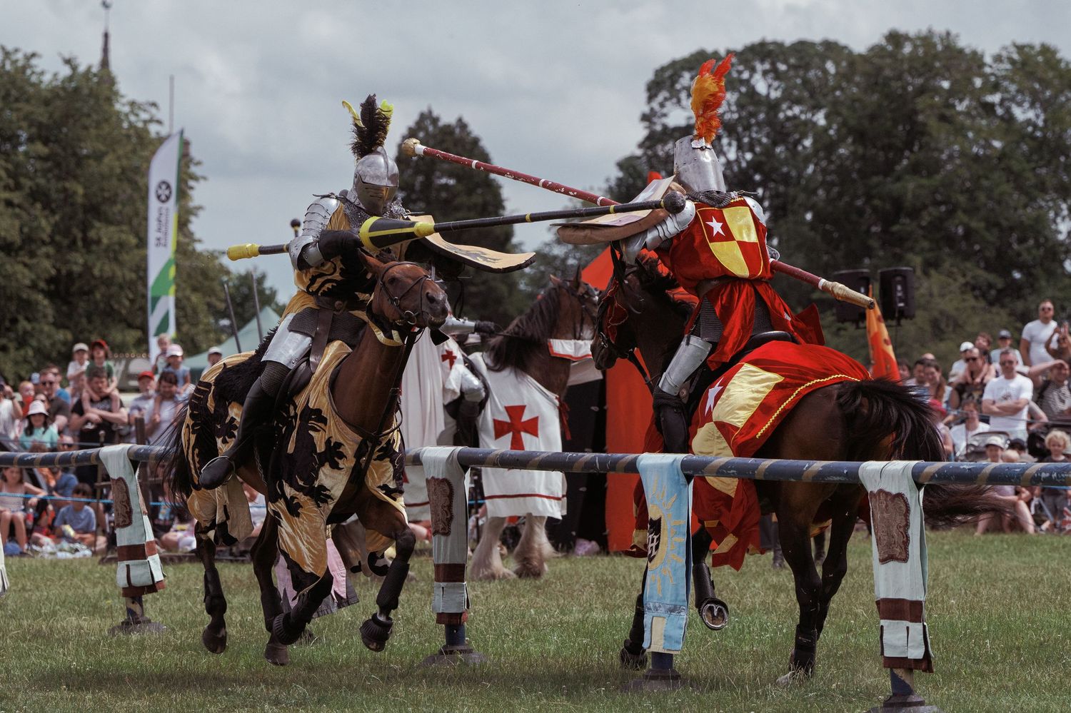 Two knights in armor jousting on horseback at a medieval reenactment event with spectators watching in the background.