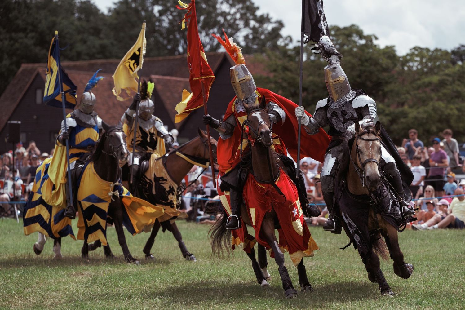 Knights in colorful armor jousting on horseback at a medieval festival reenactment on a green field.