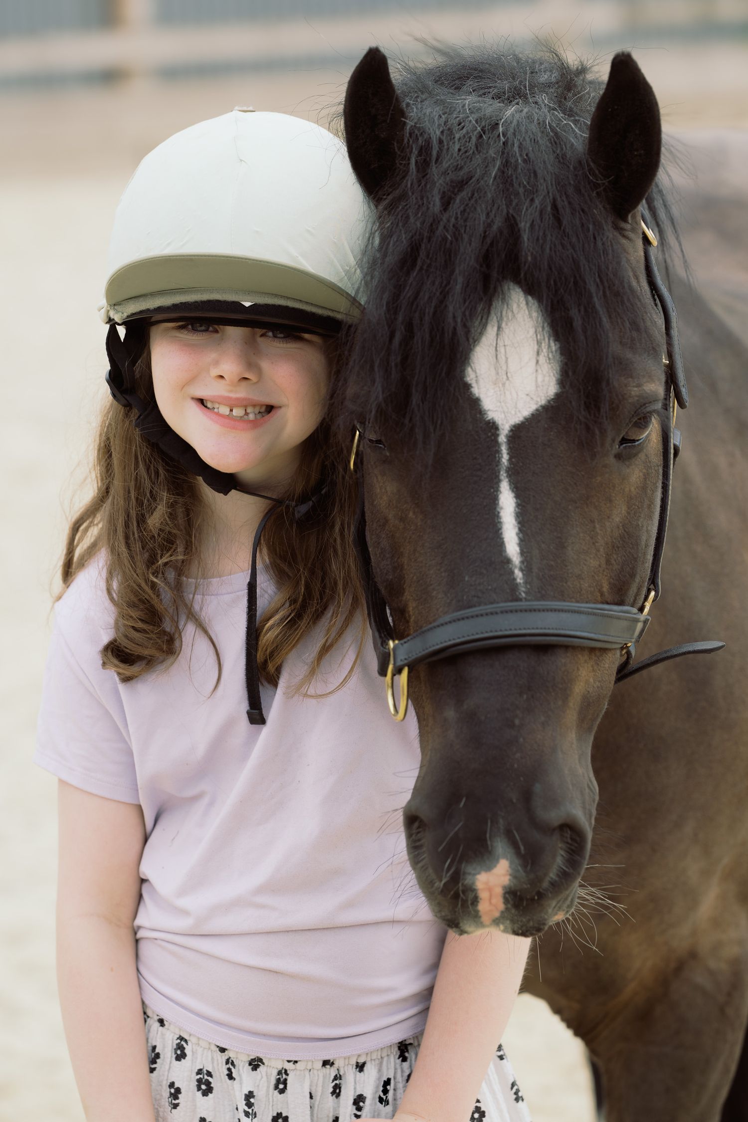 Young girl in riding helmet smiling beside a dark horse with white blaze marking in an equestrian arena.