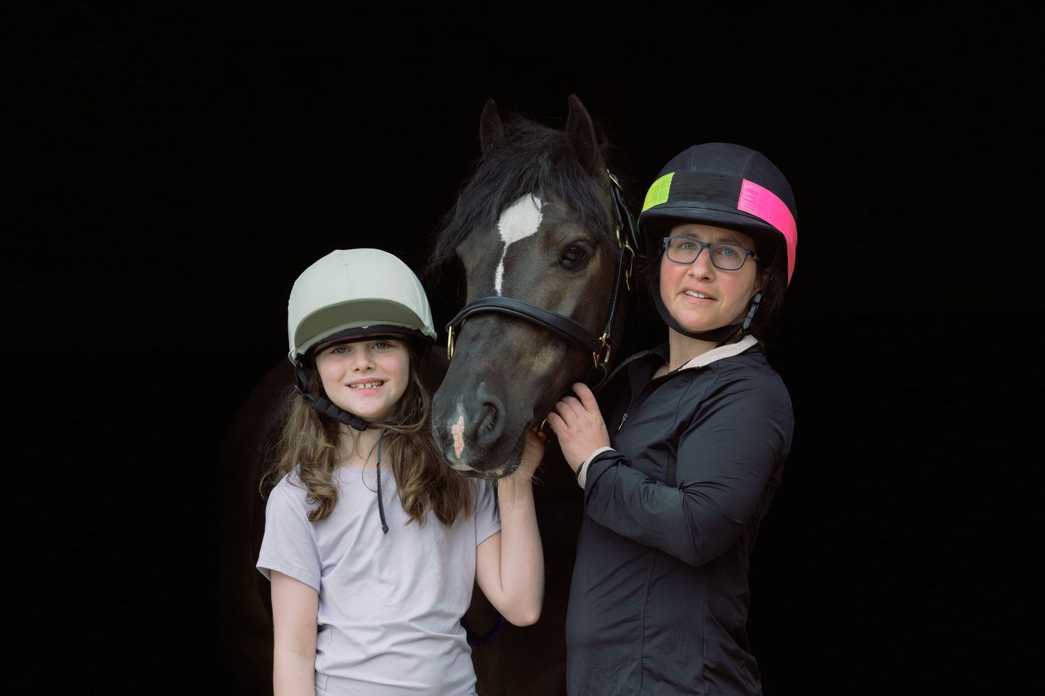 Two young equestrians wearing helmets pose with a dark horse against a black background.