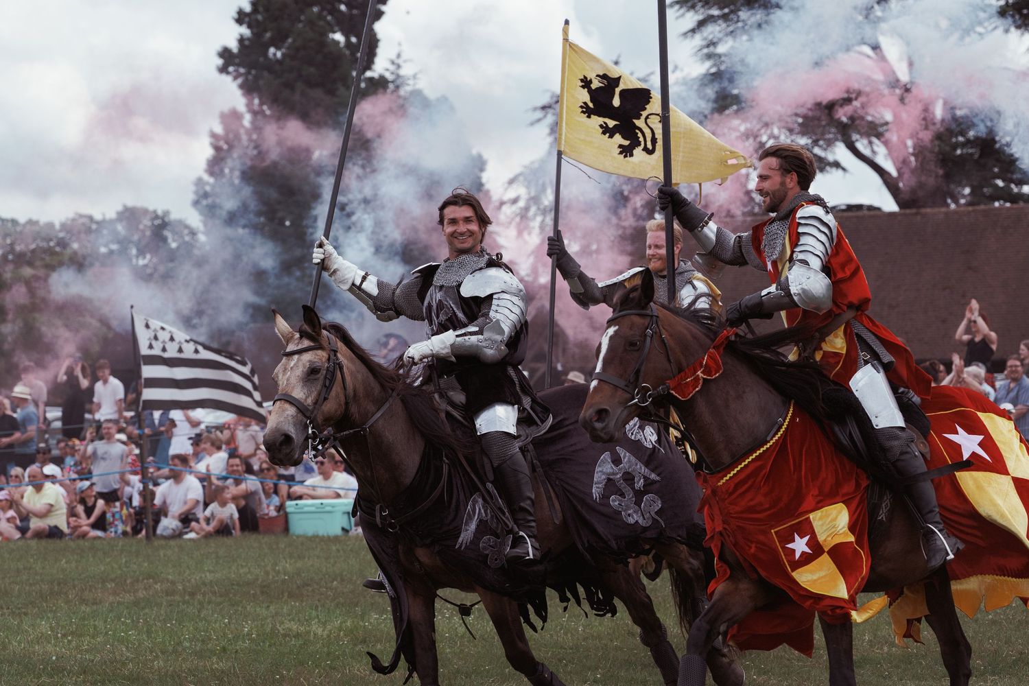 Knights in armor ride horses carrying colorful flags at a medieval jousting tournament with smoke and cheering crowd.