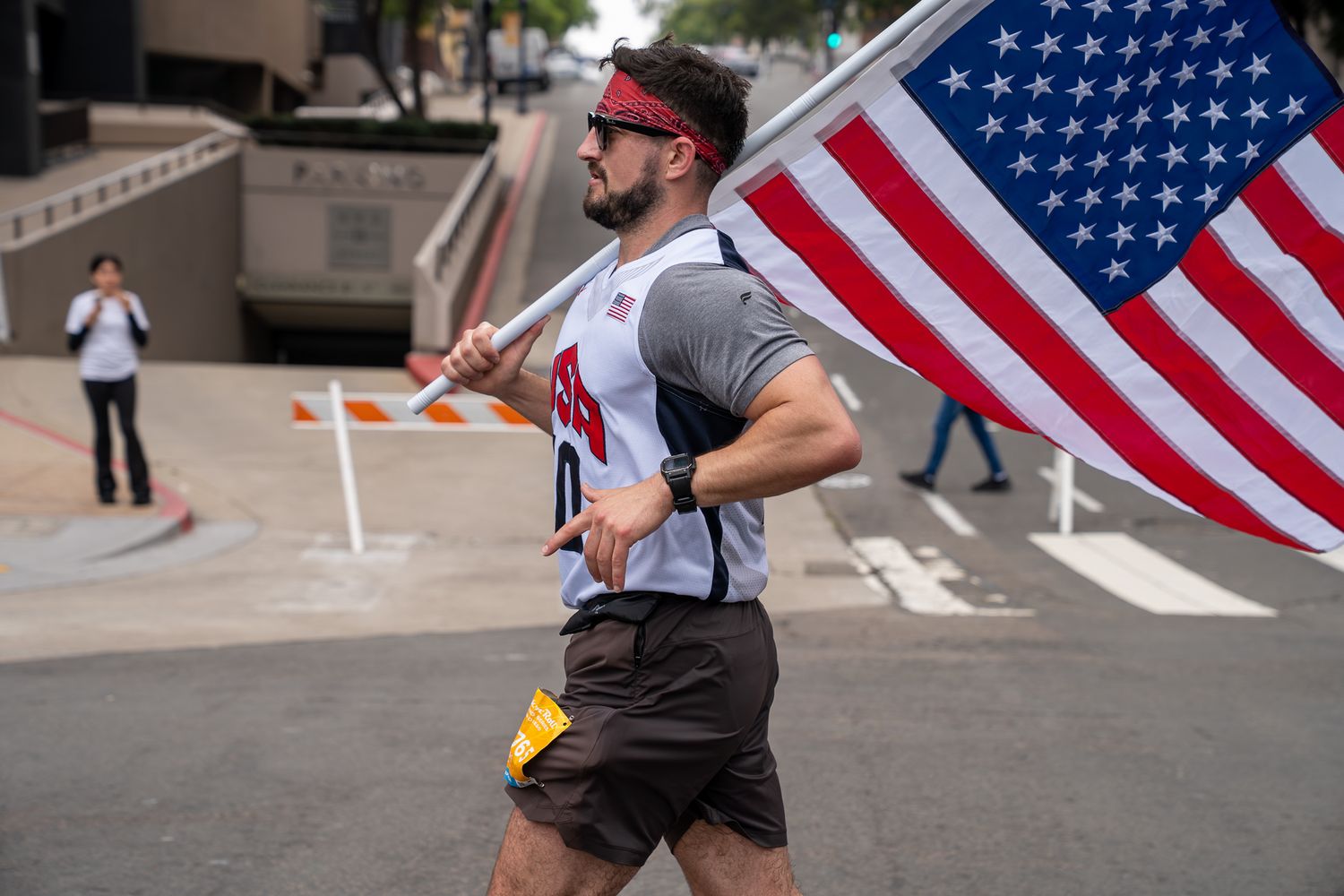 Man running with American flag wearing red headband, sunglasses, and shorts in outdoor race event.