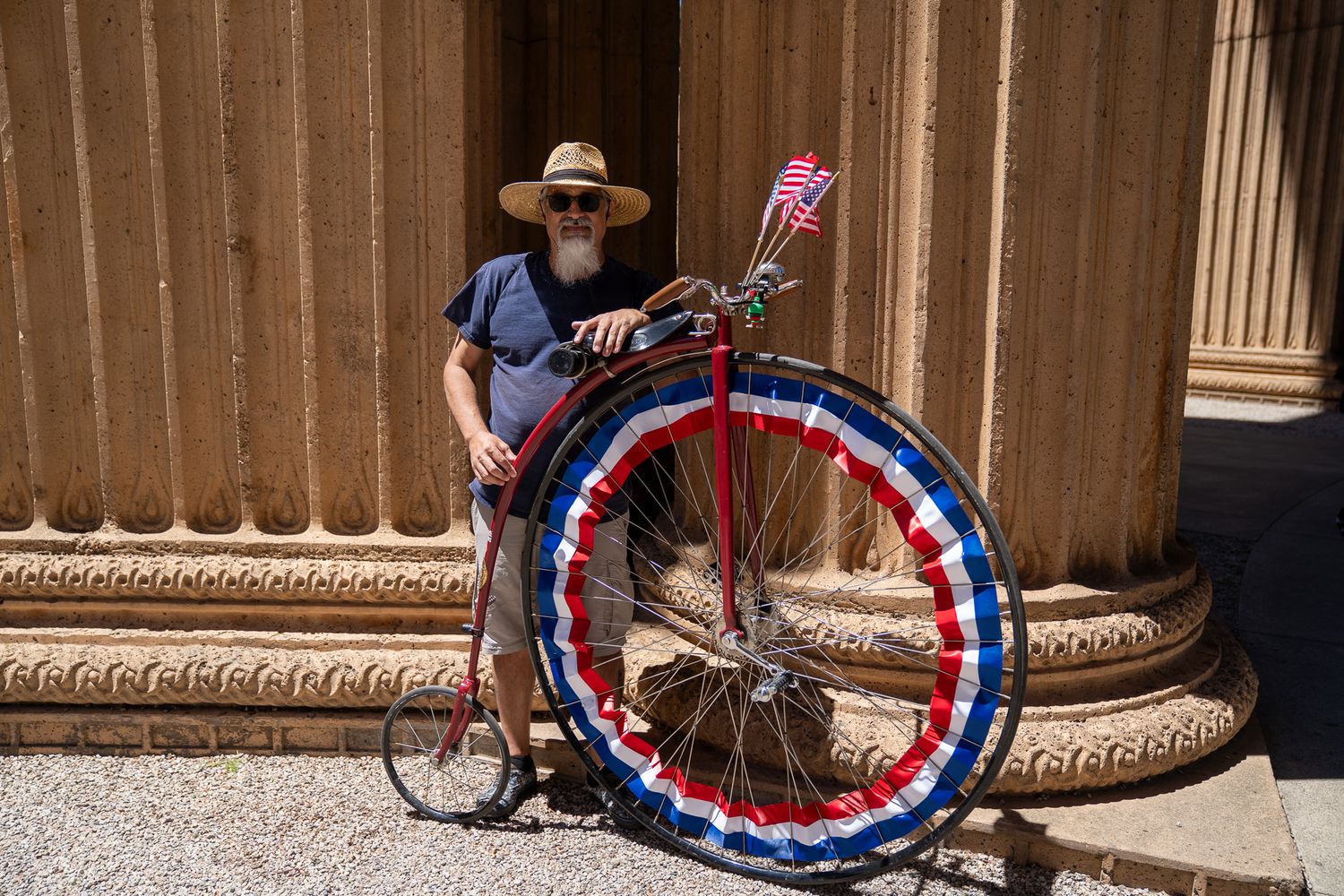 Bearded man in hat poses with patriotic penny-farthing bicycle decorated in red, white, and blue beside stone columns.