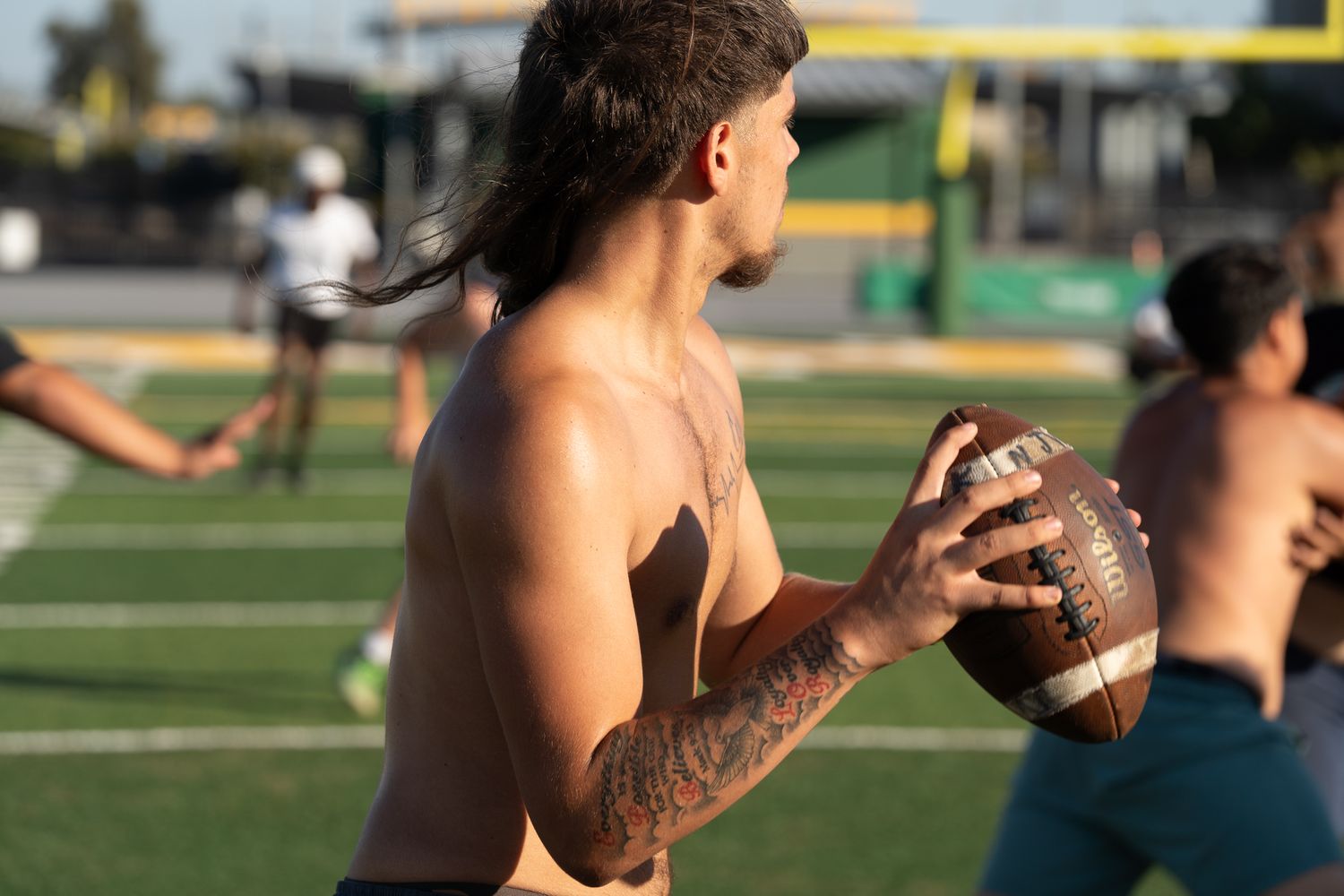 Shirtless man with tattoos and long hair holds a football on a green field during an outdoor practice game.