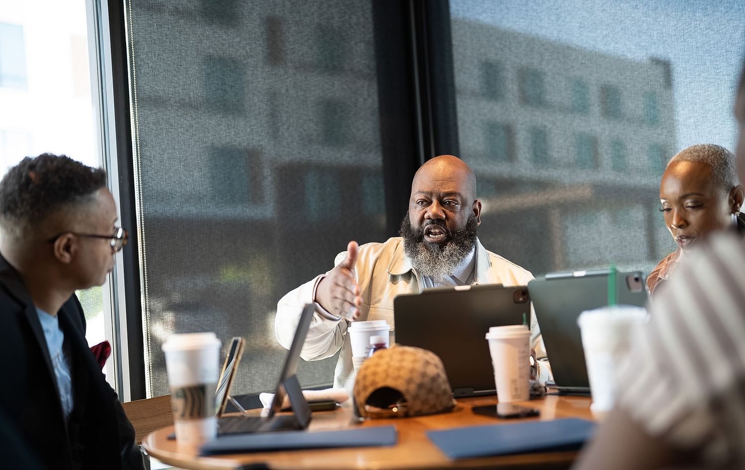 Three professionals engaged in lively discussion around a conference table with laptops and coffee cups near large windows.