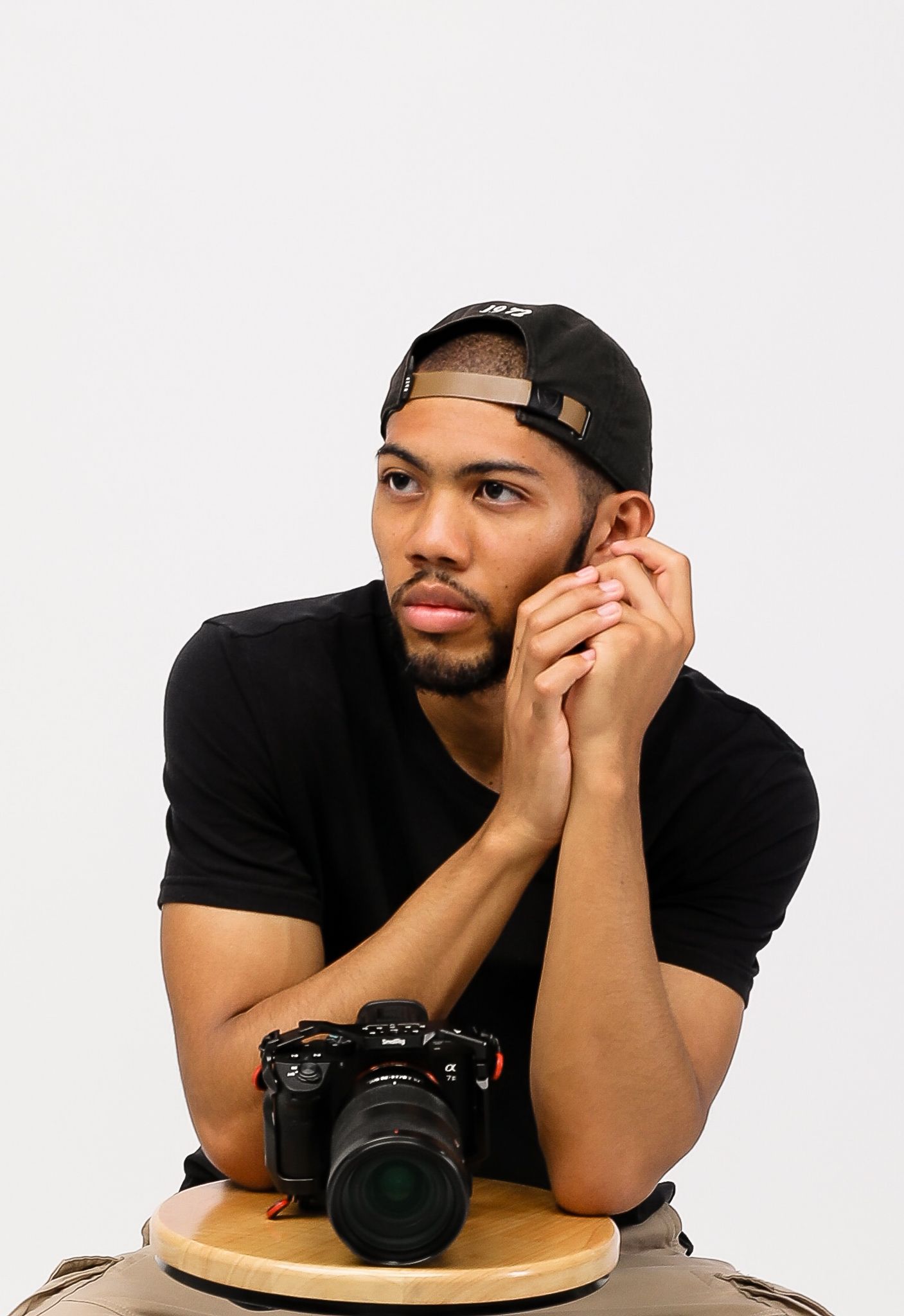 Young man in black t-shirt and snapback cap poses thoughtfully with a professional camera on a wooden surface.