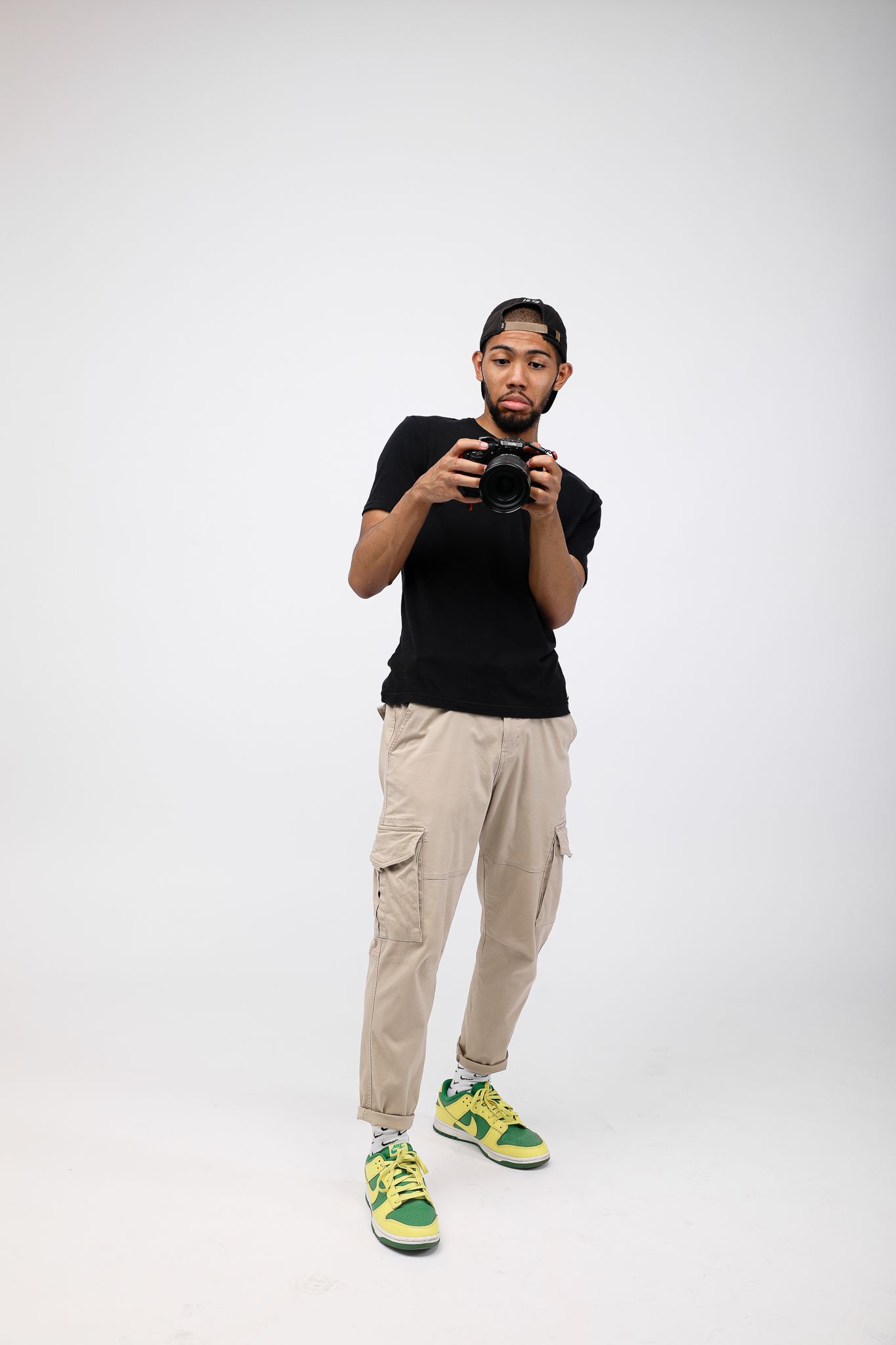 Young man in black tee, cargo pants, and yellow-green sneakers holding a camera against white background.