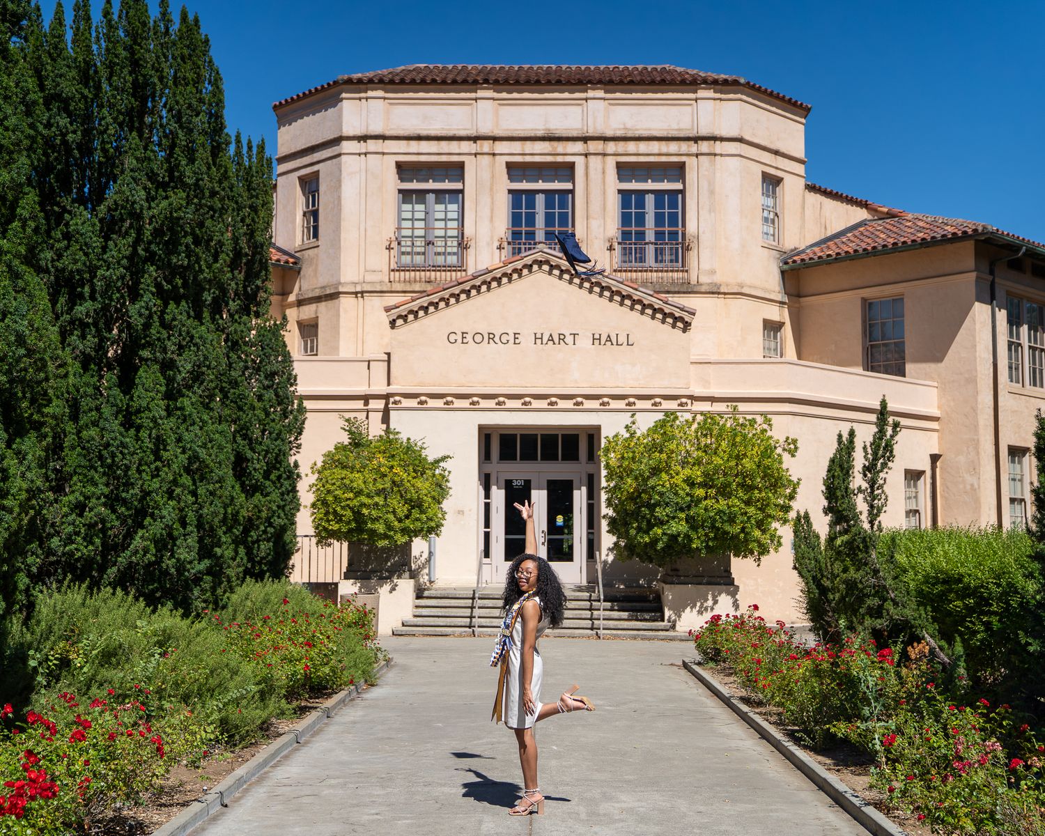 Woman posing in front of George Hart Hall building on a sunny day with red flowers and green trees.