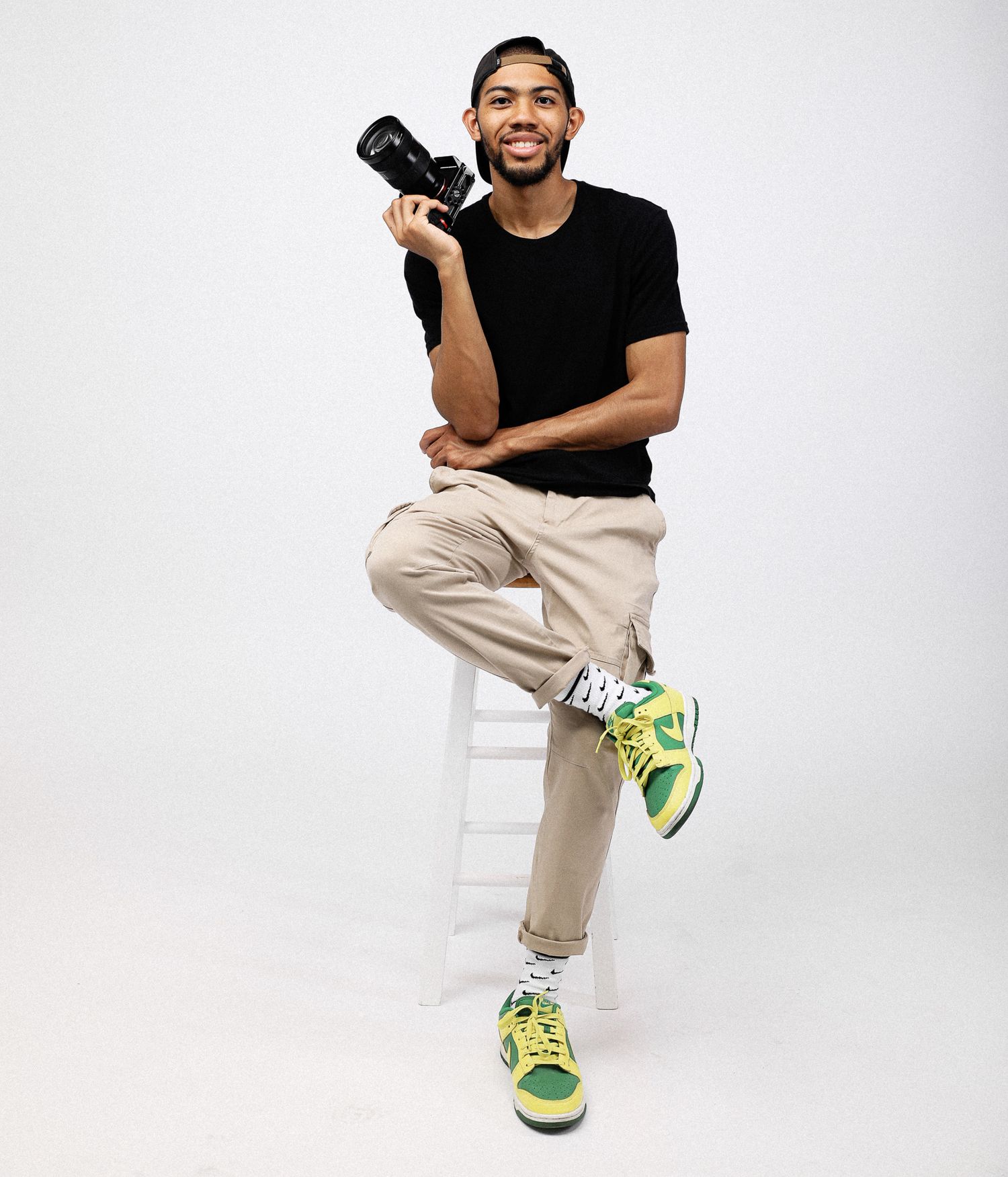 Young man sitting on stool holding camera, wearing black tee, khaki pants, and green Nike sneakers, white background.