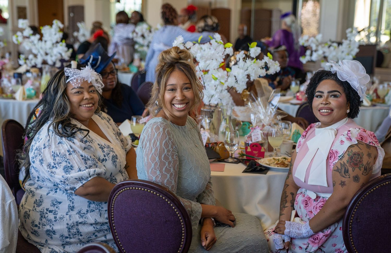 Three women in floral dresses and decorative hats smile at a elegantly decorated tea party event.