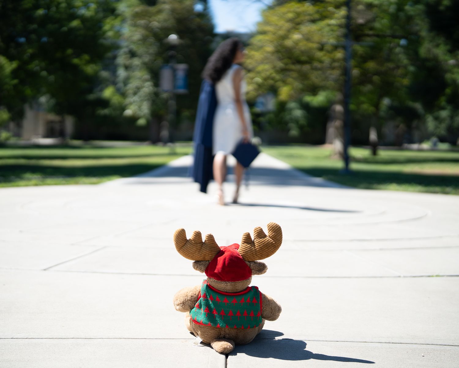 A stuffed reindeer toy sits on a sunny path as a woman walks away in the background.