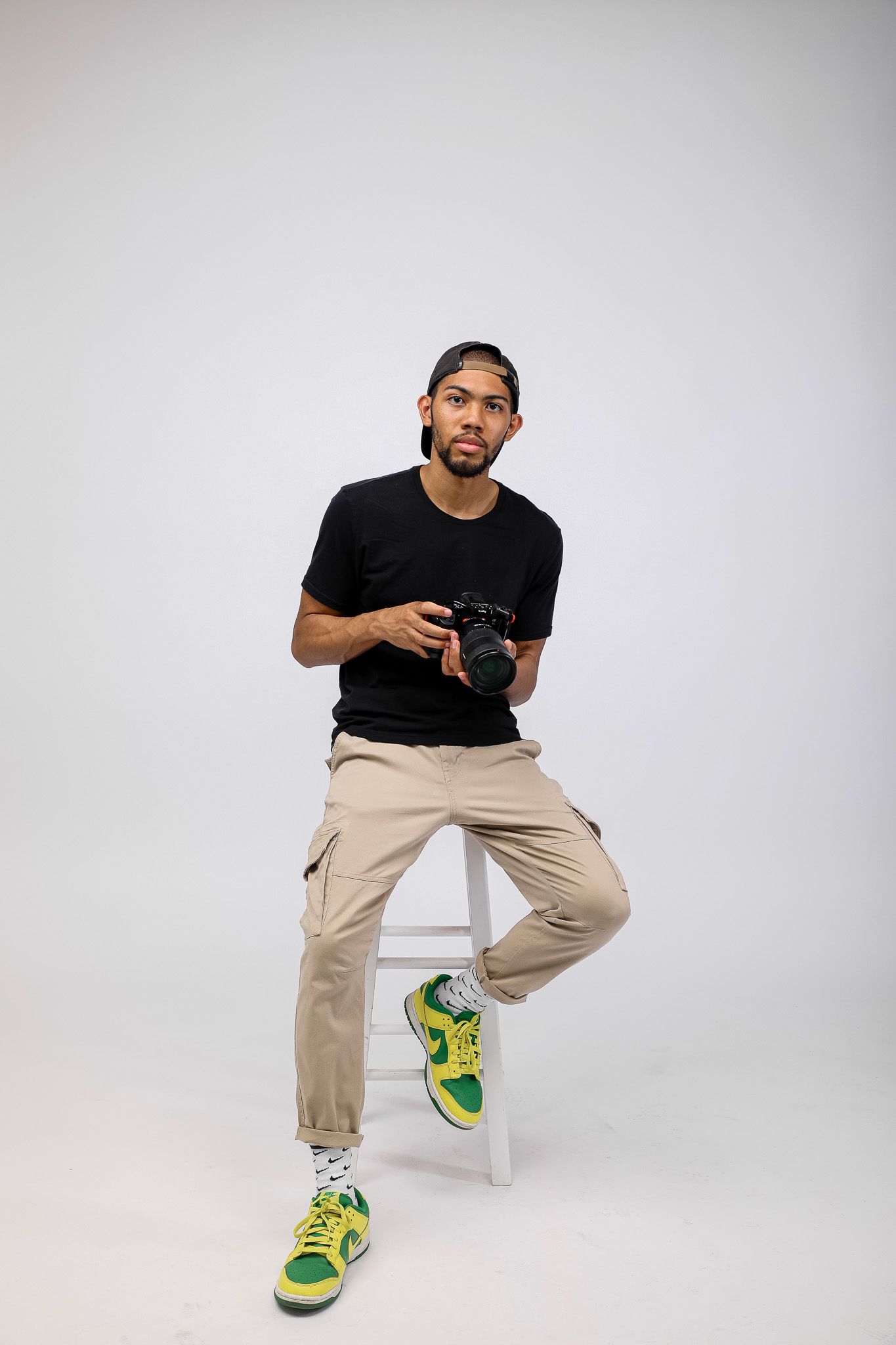 Young man in black tee, khaki cargo pants, and green sneakers sits on stool holding a DSLR camera.