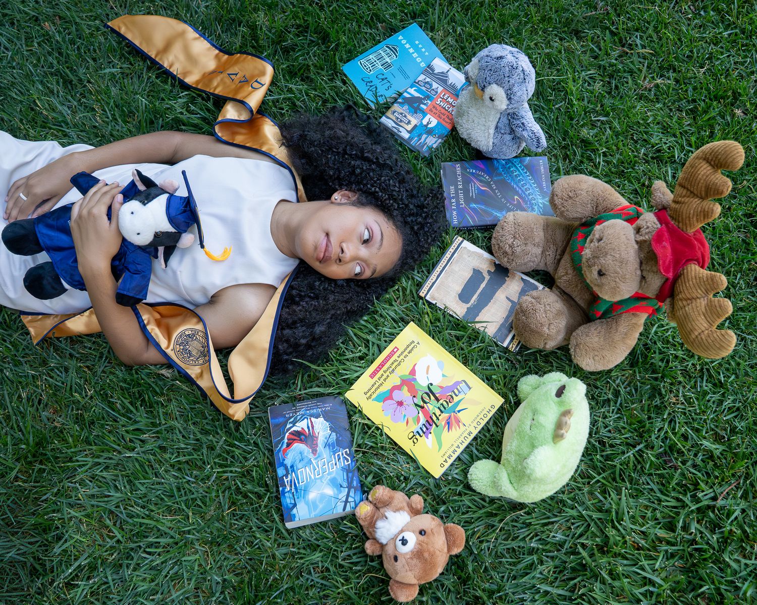 Young girl lying on grass surrounded by stuffed animals, books, and toys including a moose, elephant, and frog plushies.