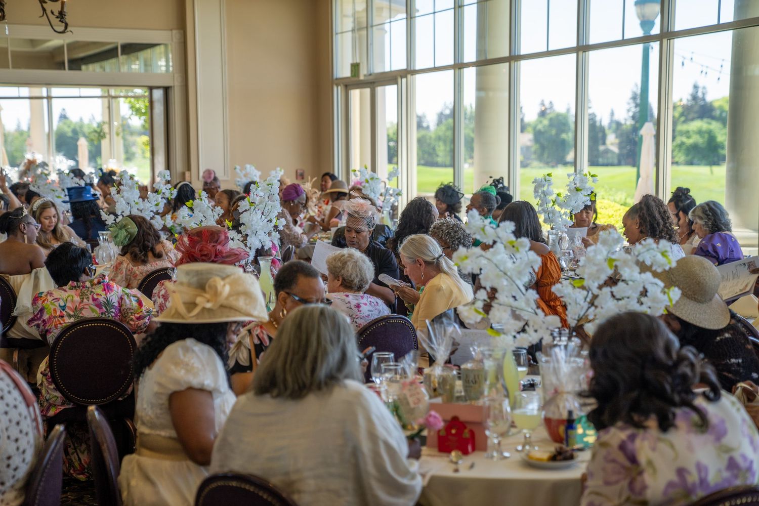 Elegant tea party luncheon with guests wearing decorative hats seated at floral centerpiece tables in a bright banquet hall.